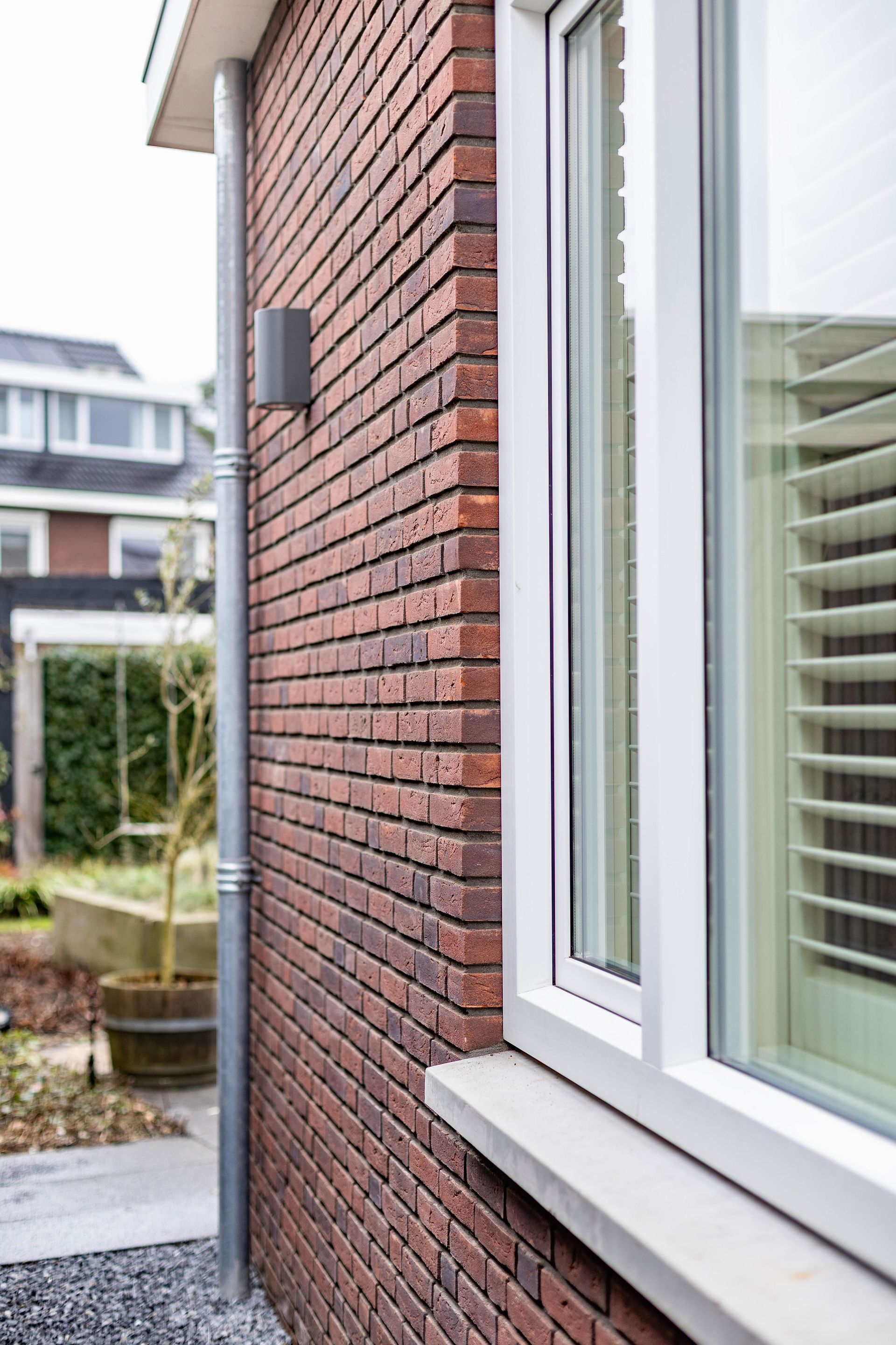 De buitenkant van een bakstenen huis met een wit omlijst raam en jaloezieën, gezien vanaf een smal zijpad.