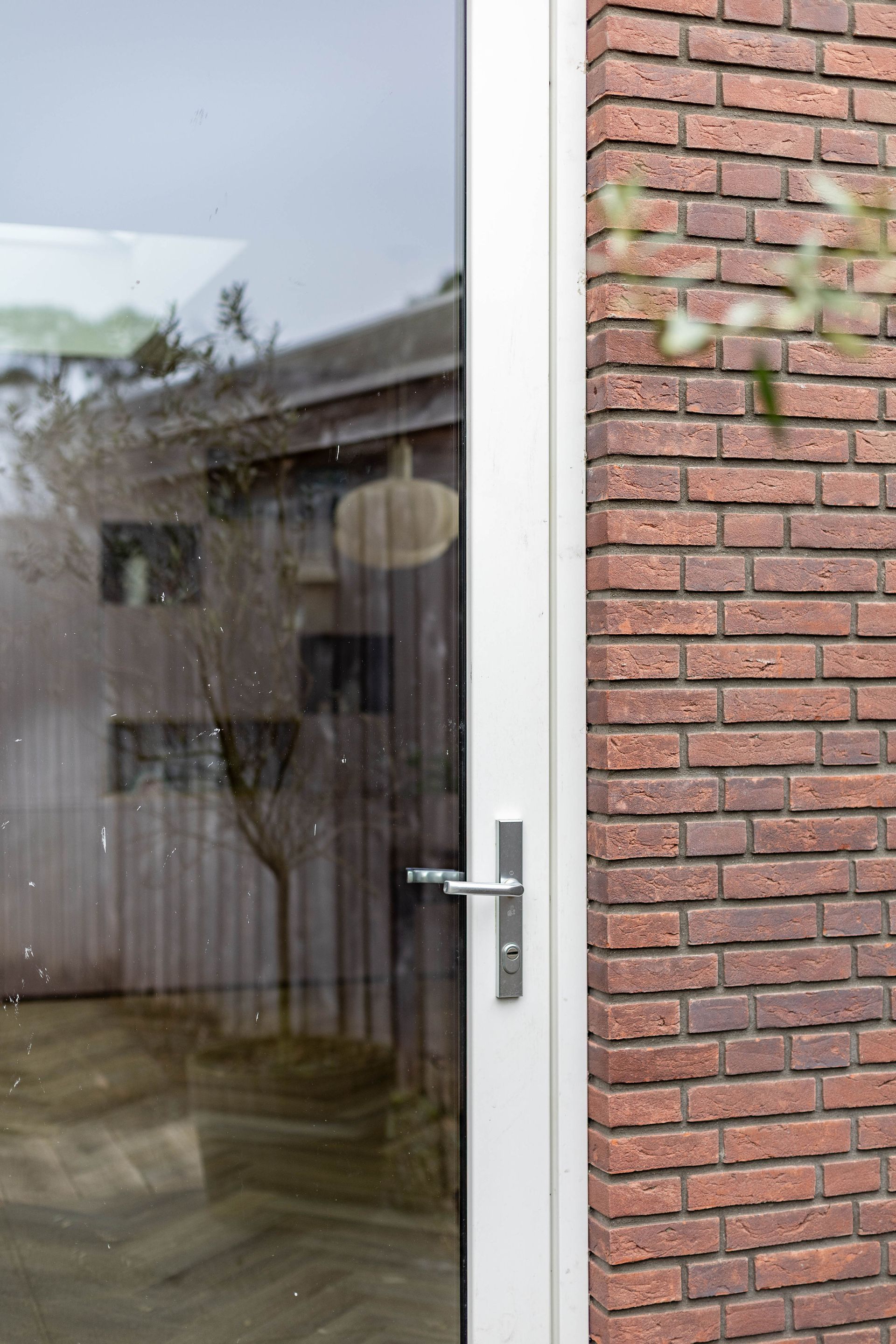 Glazen deur met wit frame naast een rode bakstenen muur, die een buitengebouw en bomen weerspiegelt.