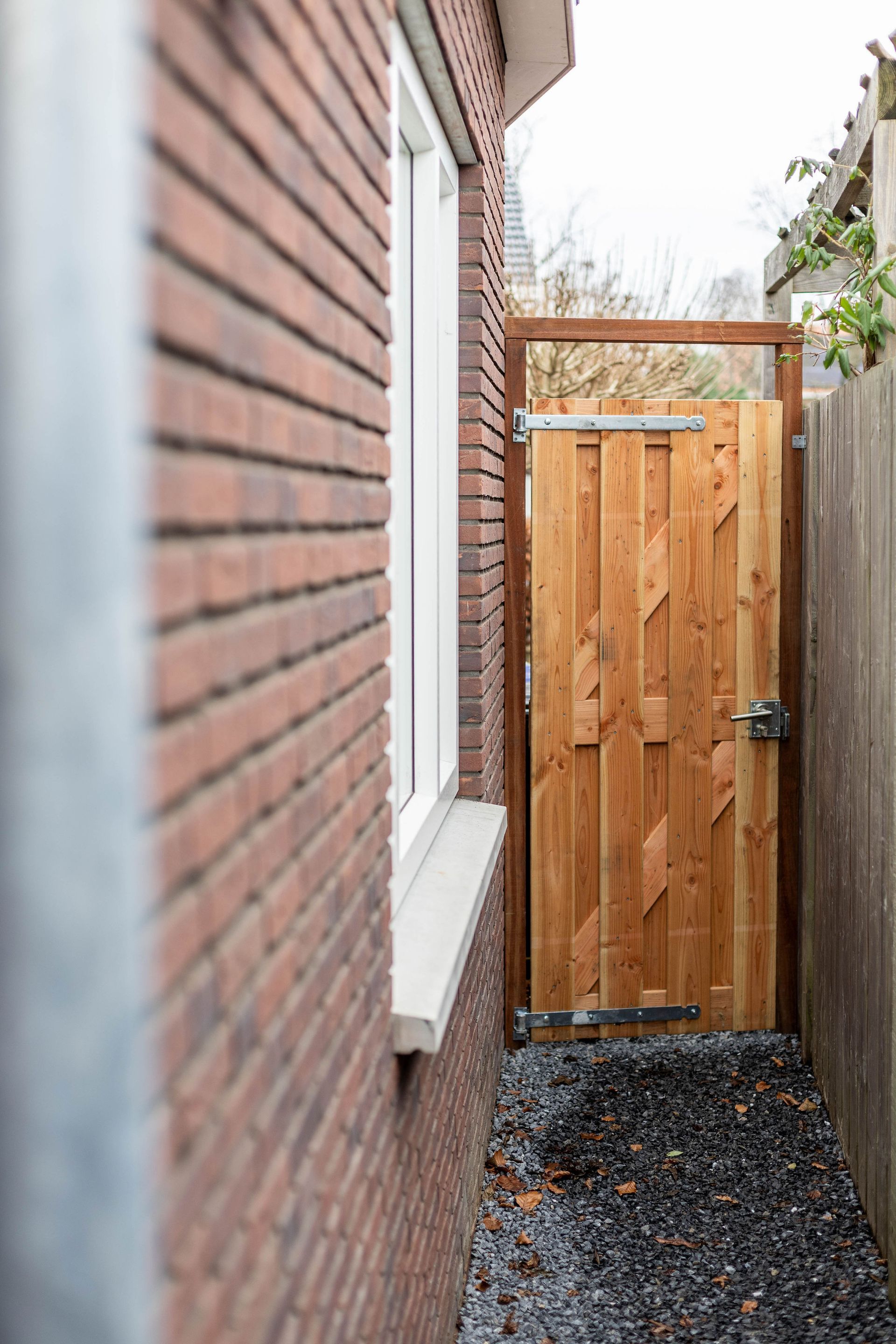 Smalle zijtuin met bakstenen muur, grindpad en een houten poort aan het einde.