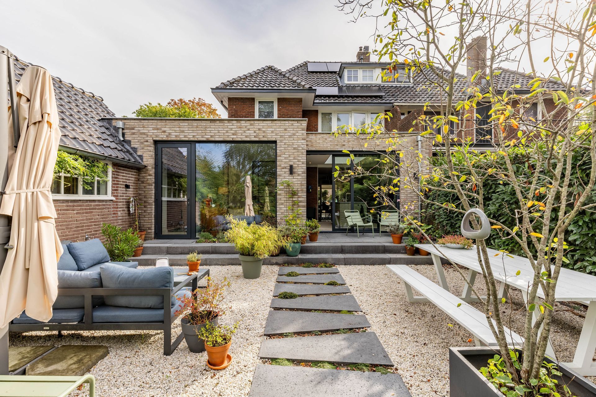 Moderne achtertuin met licht metselwerk, grote glazen schuifpuien, een terras met tuinmeubels, sierplanten en stapstenen in een grindtuin.