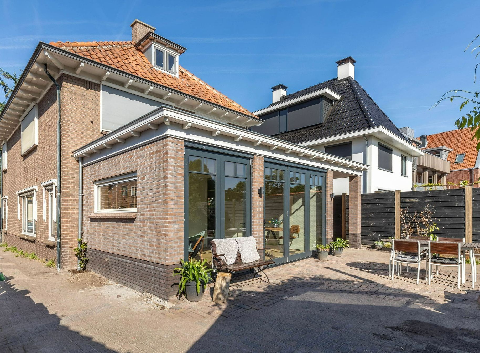 Bakstenen huis met een glazen serre en terrasstoelen in een zonnige binnenplaats.