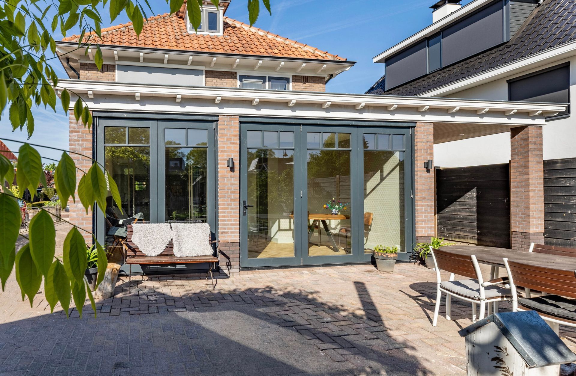 Terras in de achtertuin met glazen deuren, een witte loungestoel en een bakstenen gevel in het felle zonlicht.