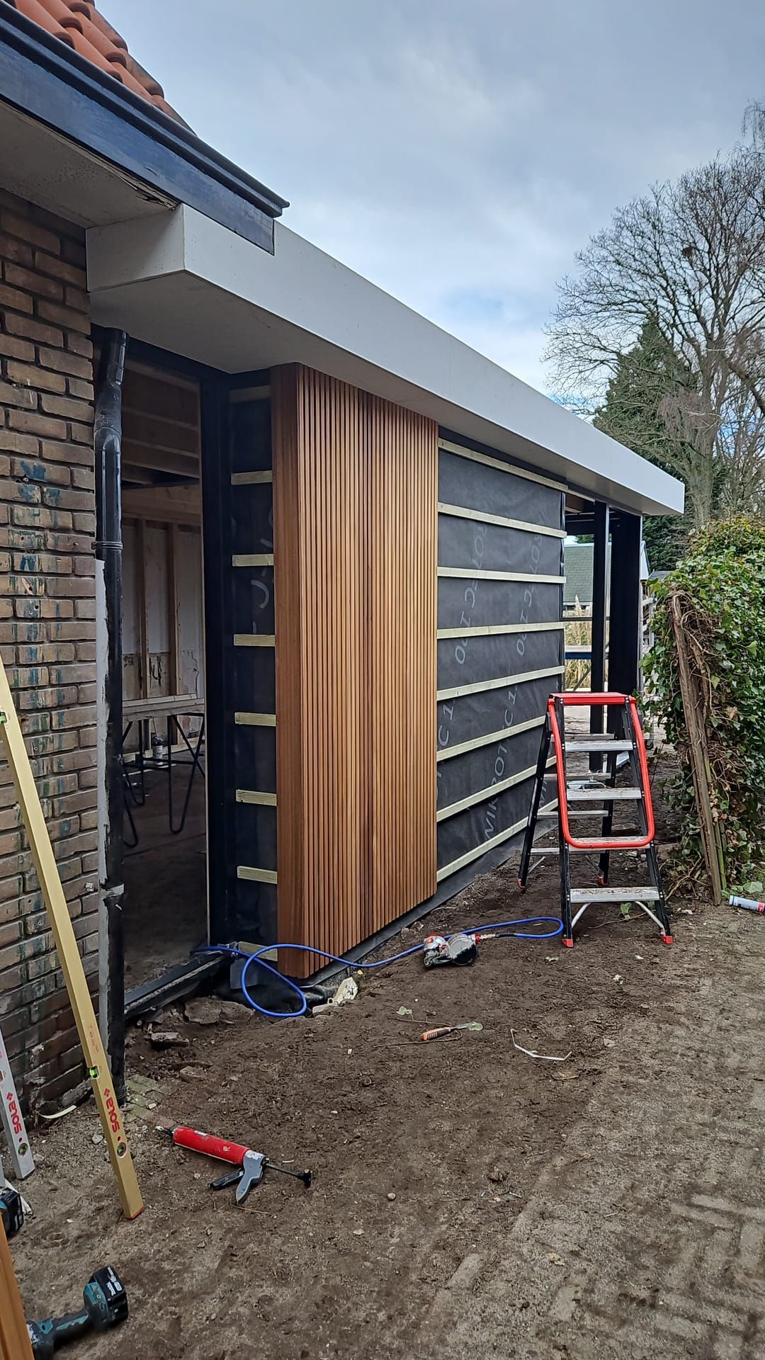 De buitenkant van een huis in aanbouw, met houten gevelbekleding, een ladder en gereedschap naast een garagedeur.