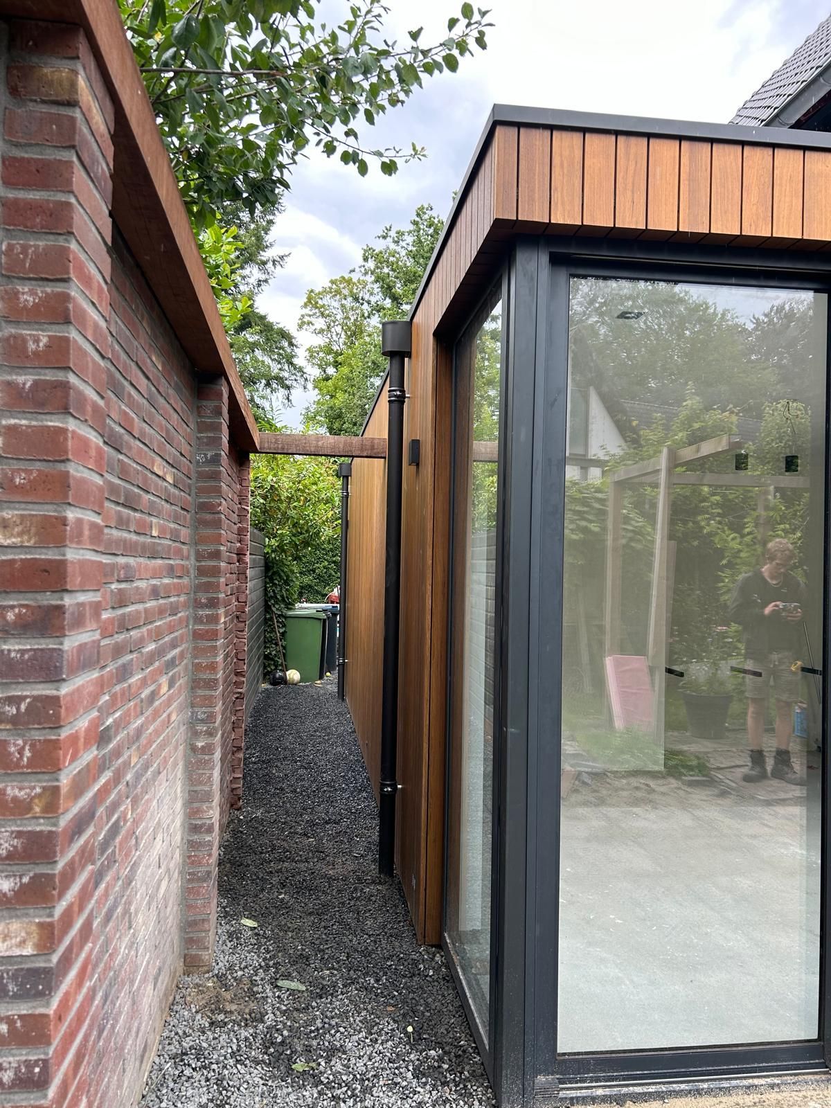 Smalle grinddoorgang tussen een bakstenen muur en een modern gebouw van glas en hout.