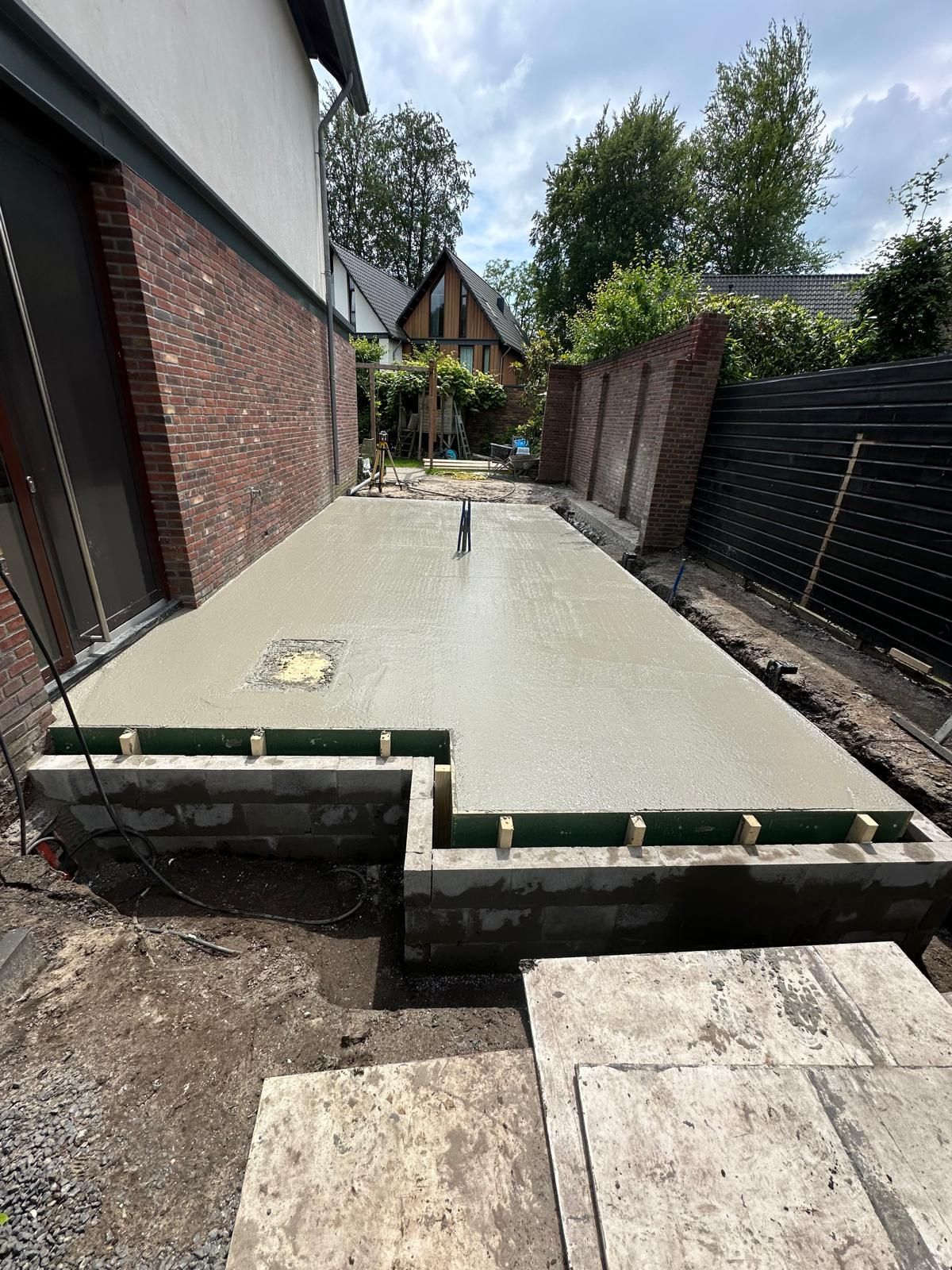 Een nieuw betonnen terras wordt aangelegd naast een bakstenen huis met een omheinde tuin.