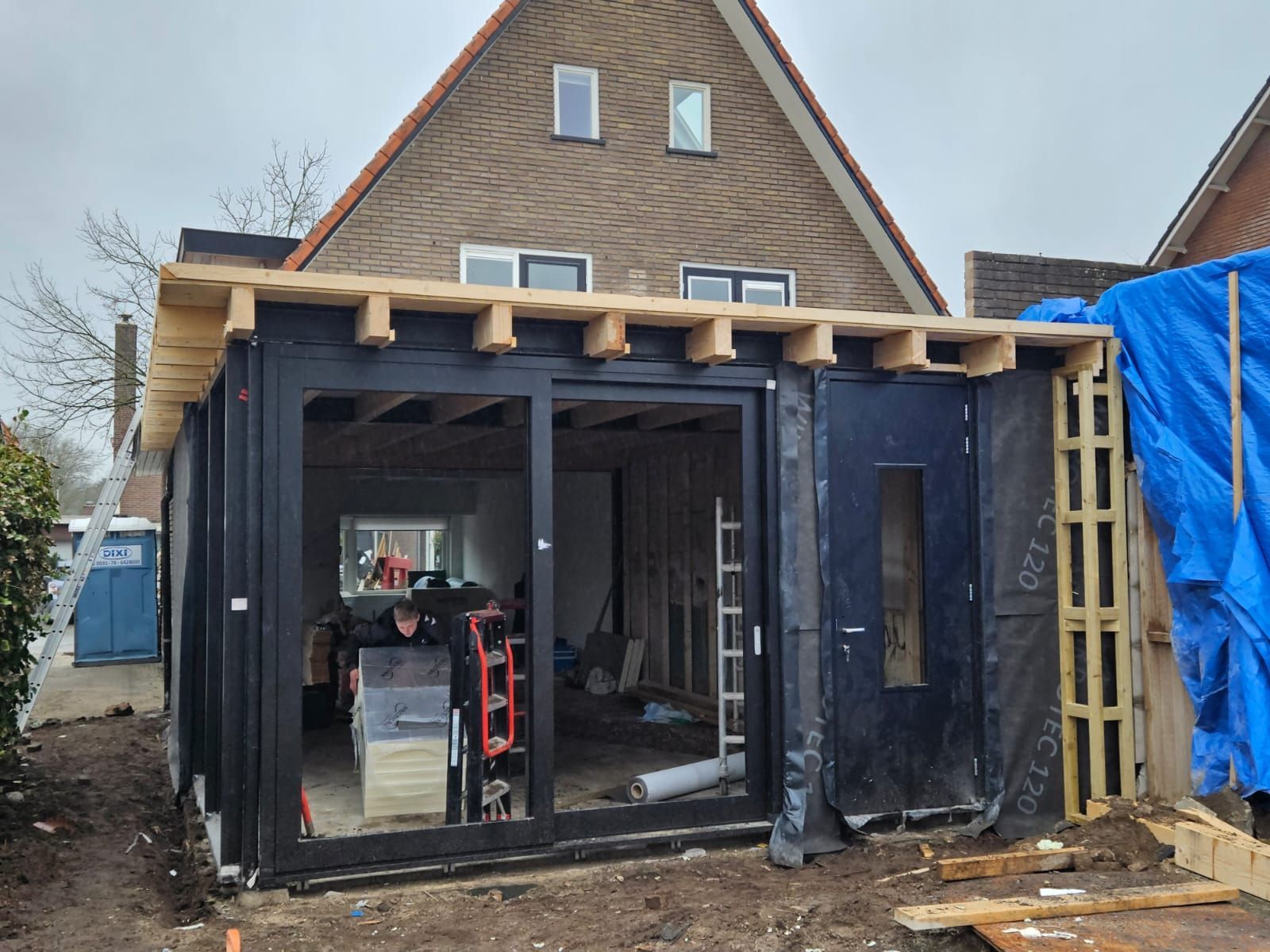 Gedeeltelijk gebouwde aanbouw met zwart omlijste opening, zichtbare dakbalken en bouwmaterialen aan de binnenkant.