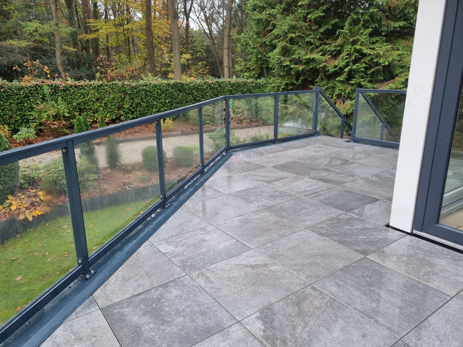 Terras met grijze tegels en glazen leuning. Terrasrenovatie en verbouwing buitenruimte.