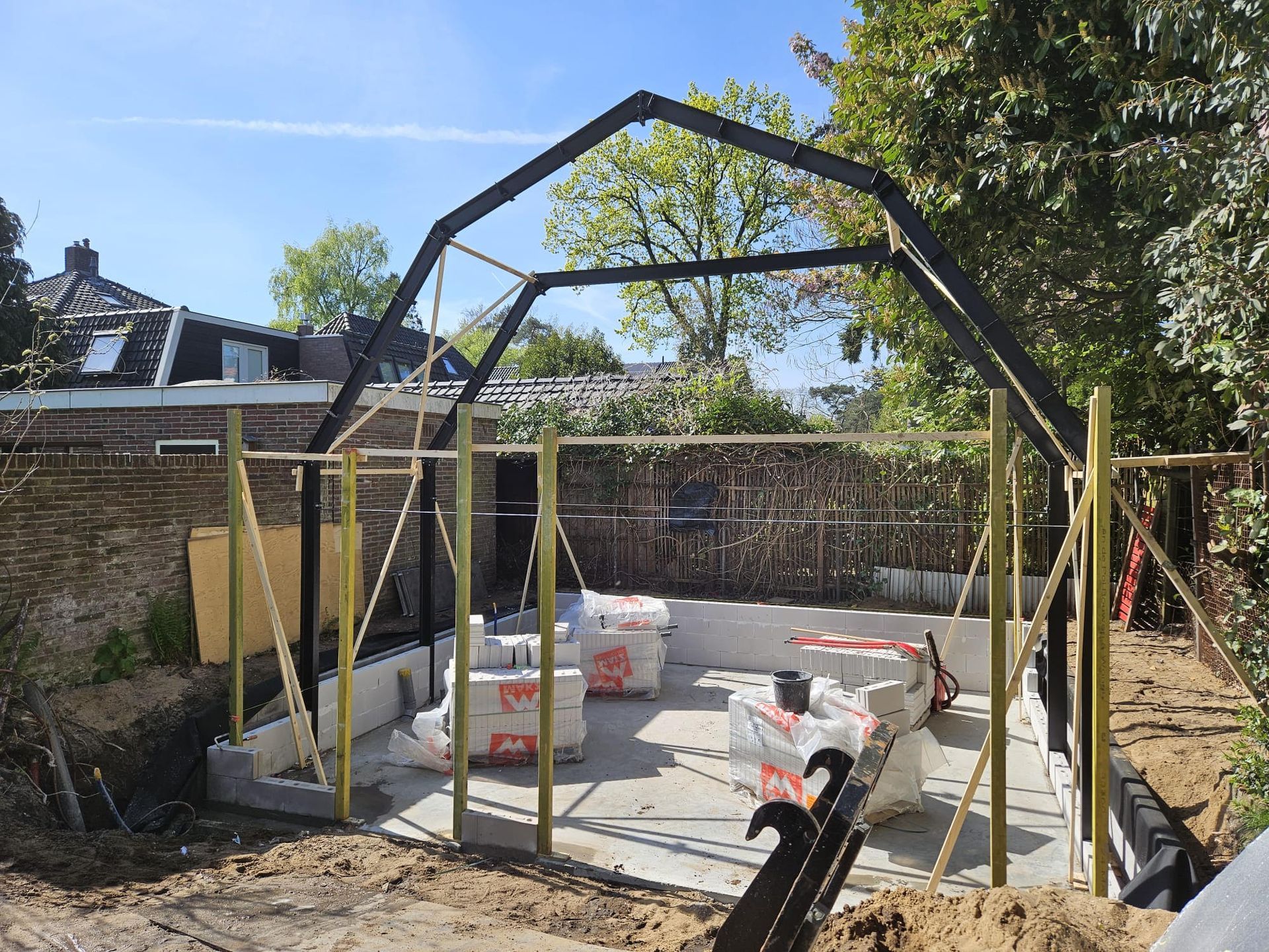 Gedeeltelijk gebouwde stalen schuilplaats op een betonnen fundering met bouwmaterialen in een achtertuin.
