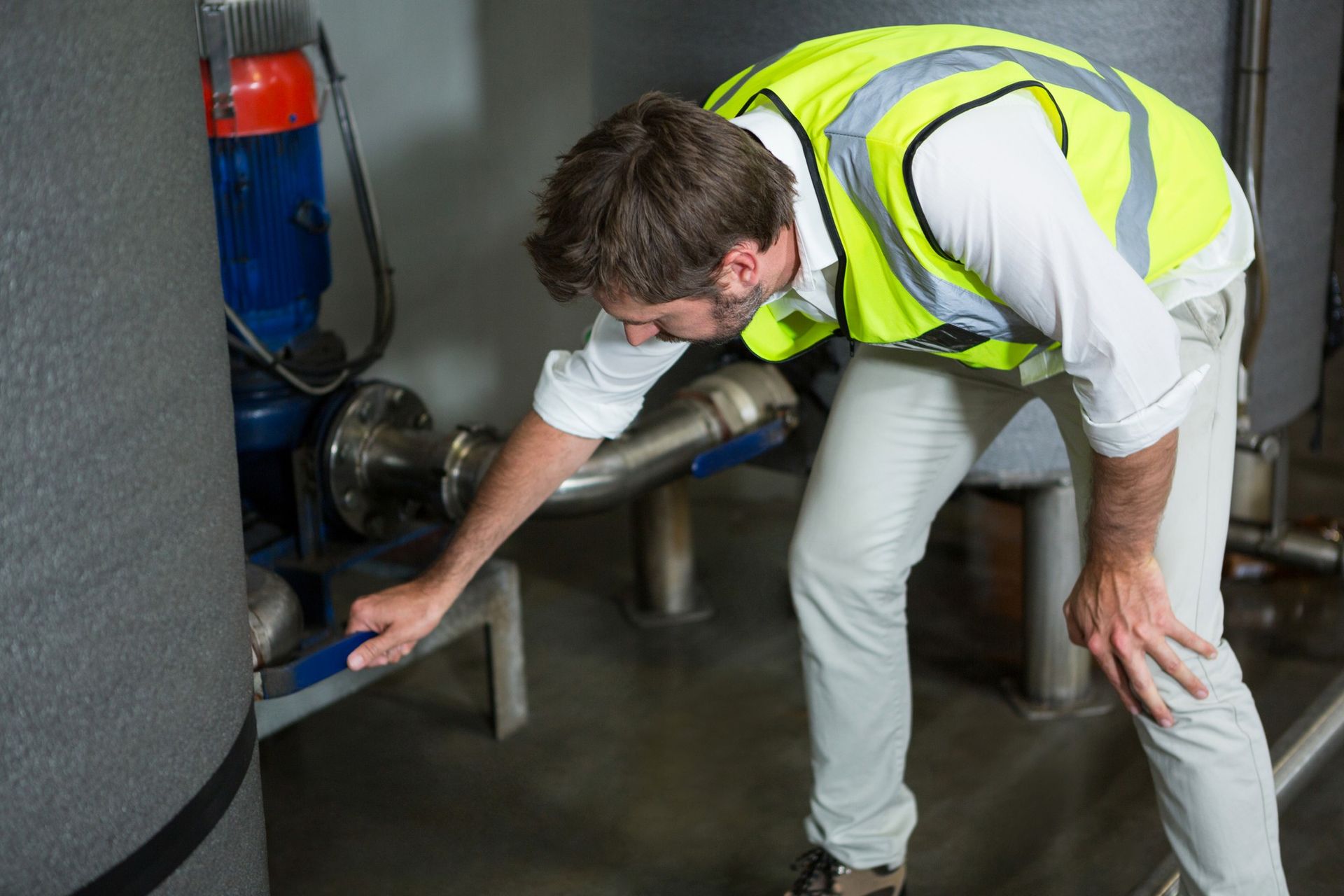A man in a yellow vest is inspecting a pipe in a factory.