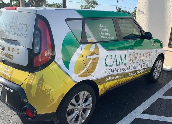 Wrapped compact car parked in a lot, advertising CAM Realty Commercial Services in green, yellow, and white.