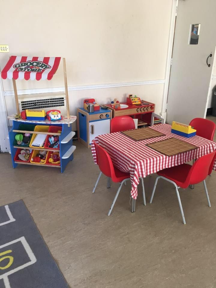 Play kitchen area: toy stand, table with checkered tablecloth, red chairs, and other play food toys.