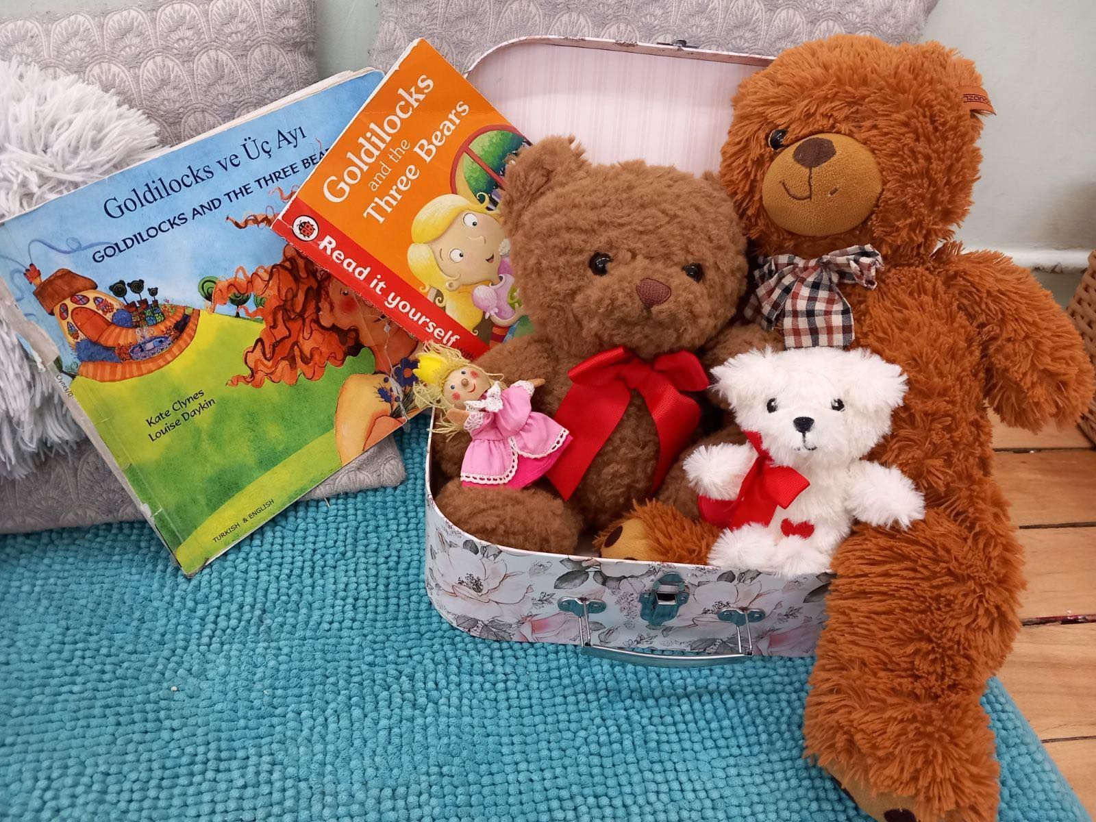 Teddy bears and storybooks in a floral-patterned container, set on a blue rug, with a soft blanket in the background.