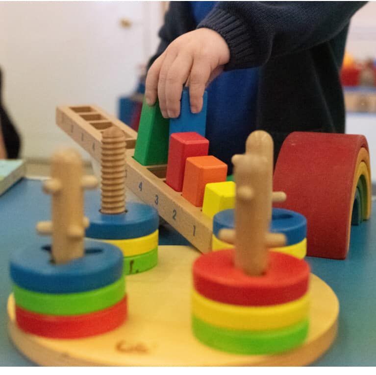 Child's hand reaching for a green block on a wooden toy with colorful rings and blocks on a table.