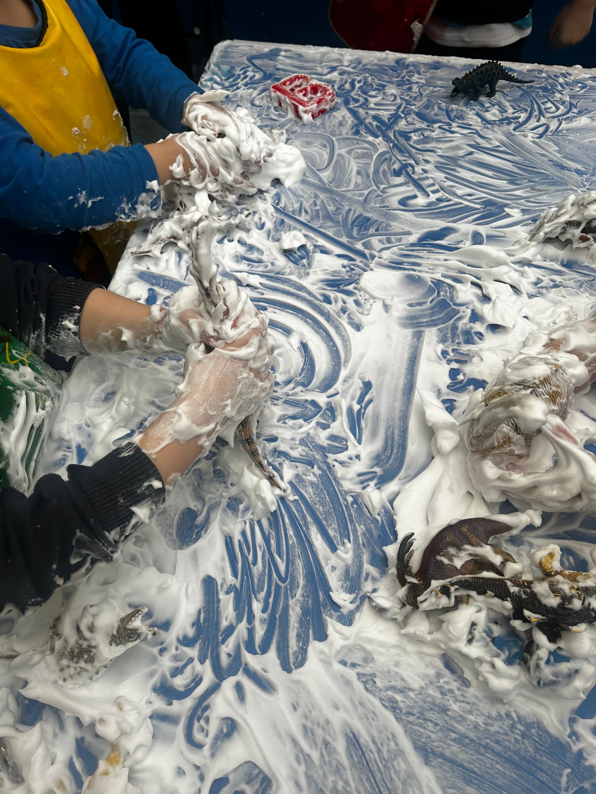 Children's hands covered in white foam, playing and smearing on a blue table.