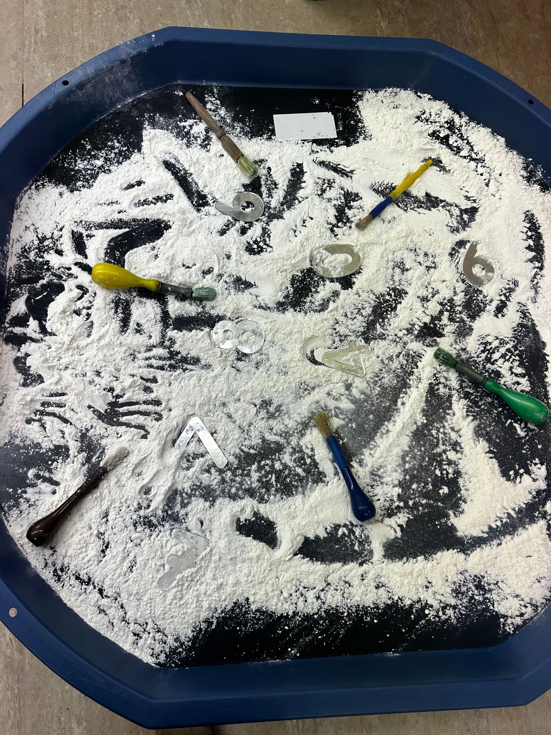 A blue tray filled with white powder and tools. Handprints and shapes are etched into the powder.