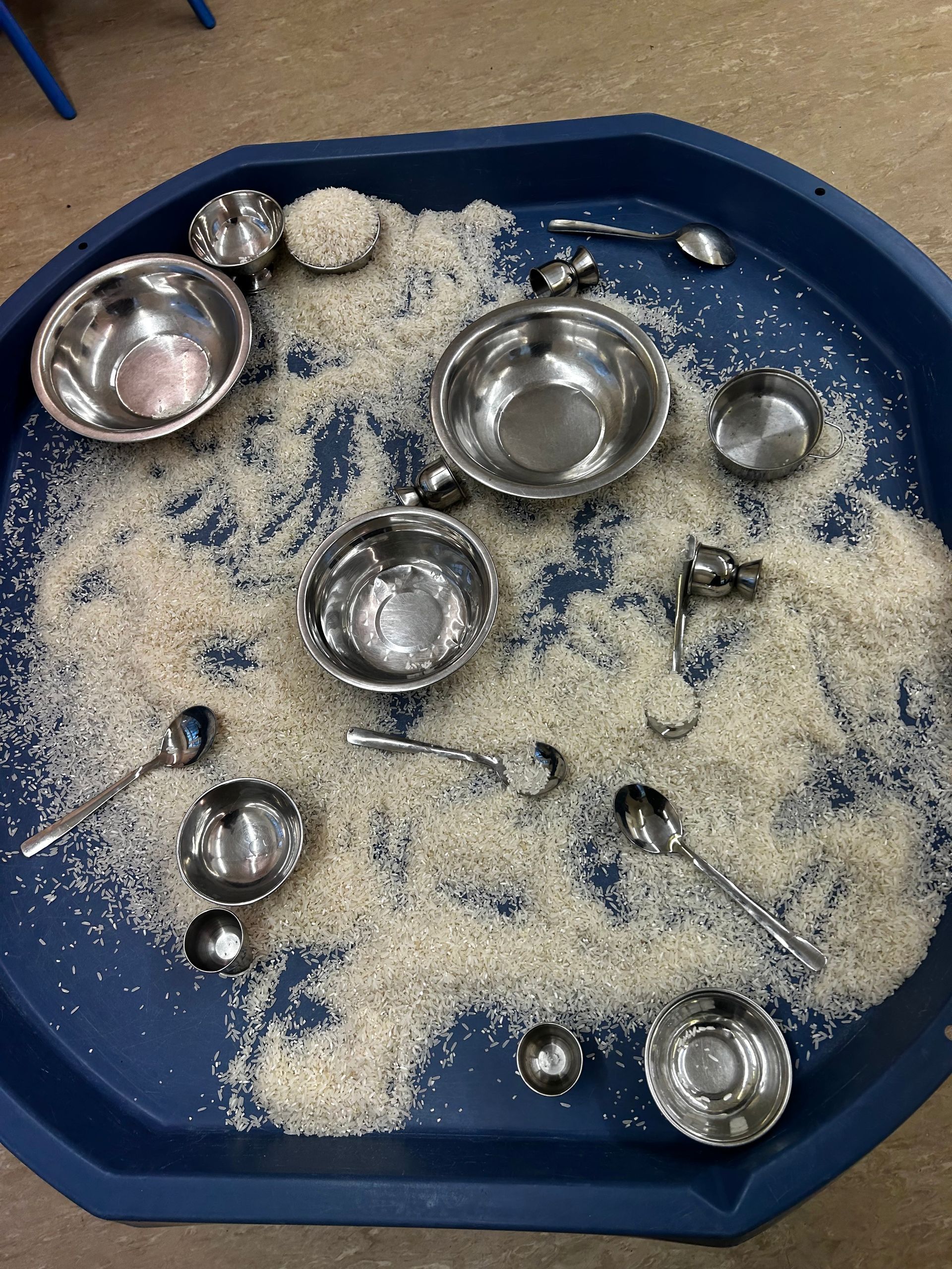 Blue tray filled with rice, assorted metal bowls and spoons for sensory play.