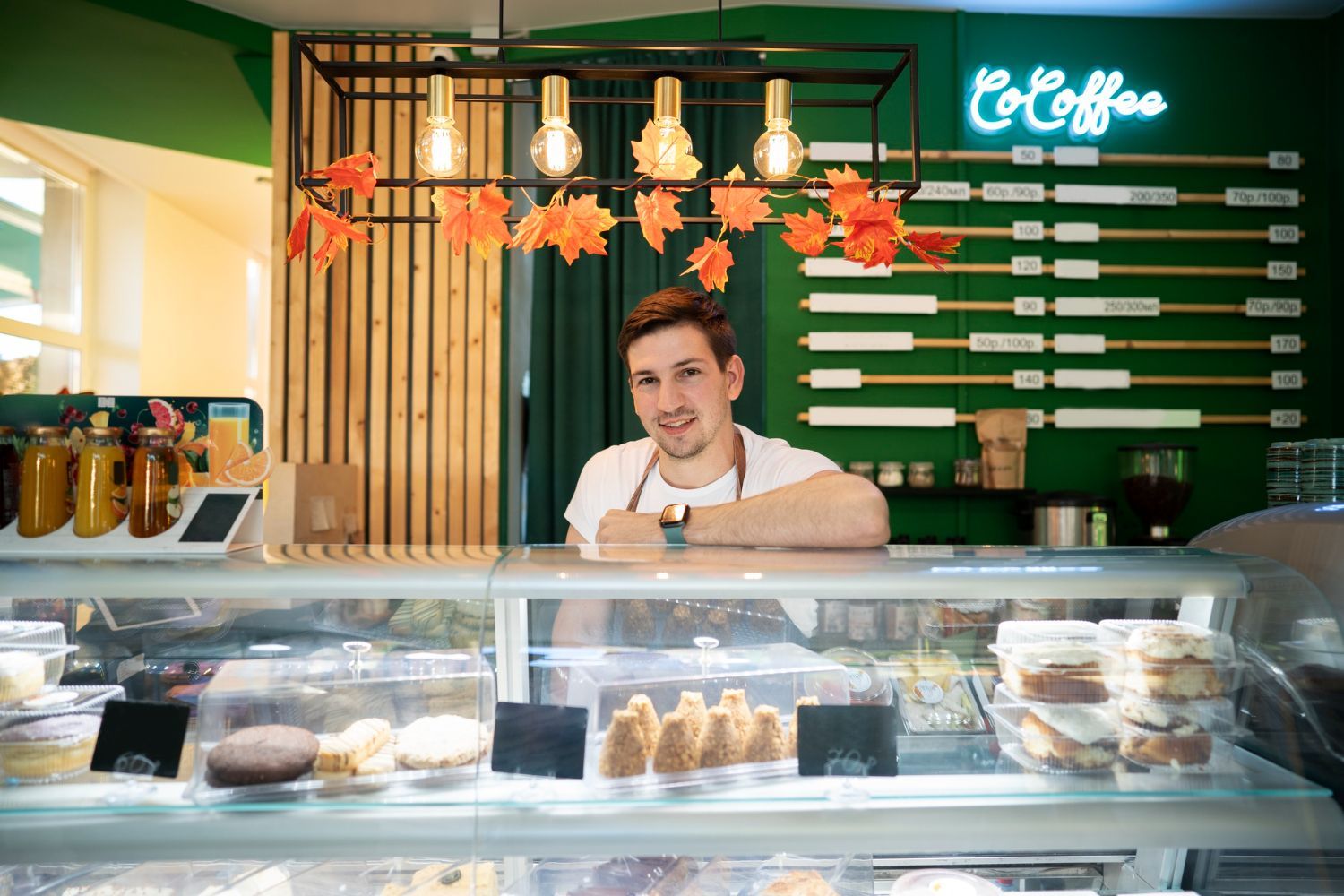 Smiling barista behind a bakery counter with pastries and hanging autumn leaves