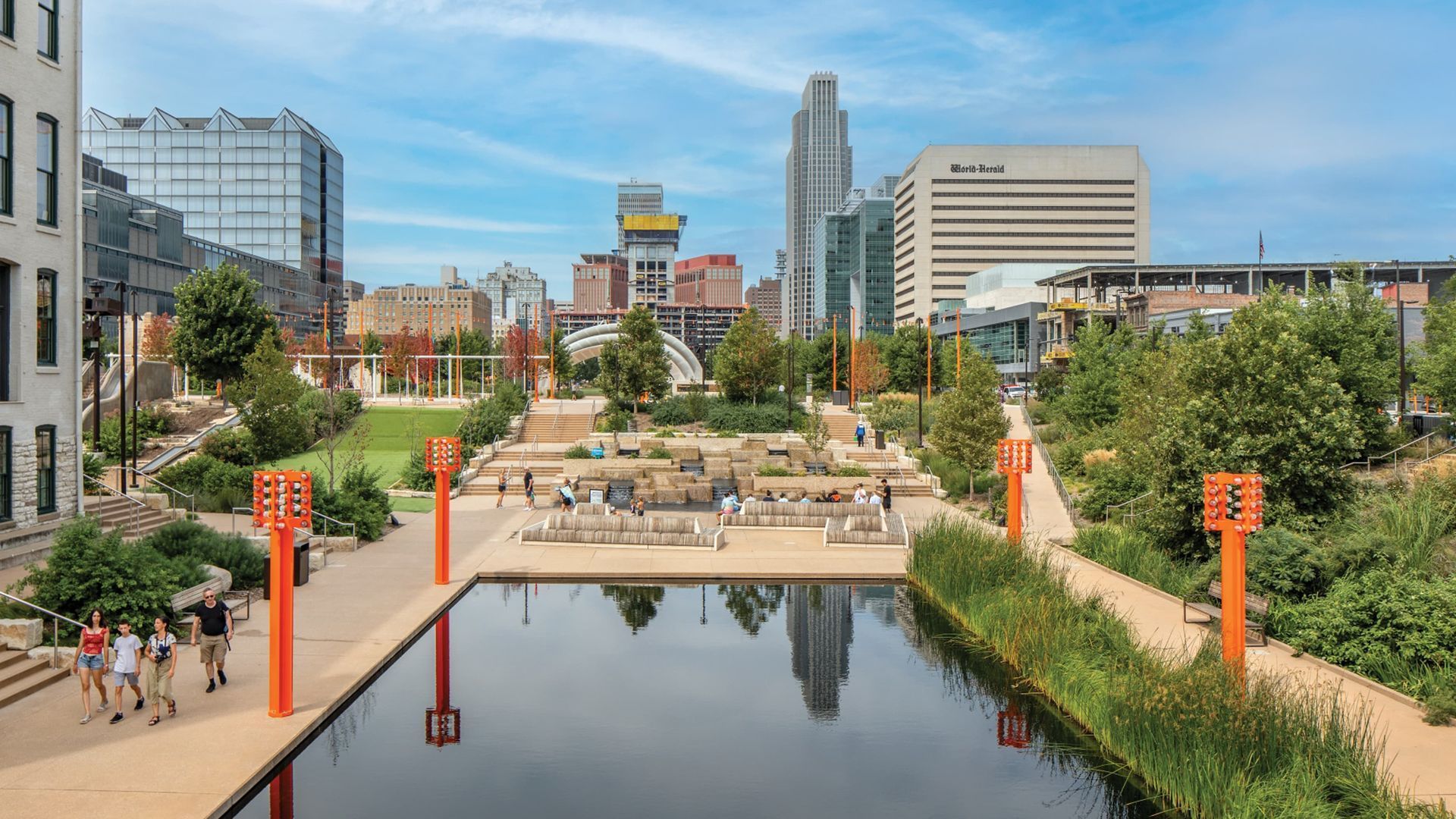 Urban canal park with a reflecting pool, orange lamp posts, people walking, and city buildings in the background