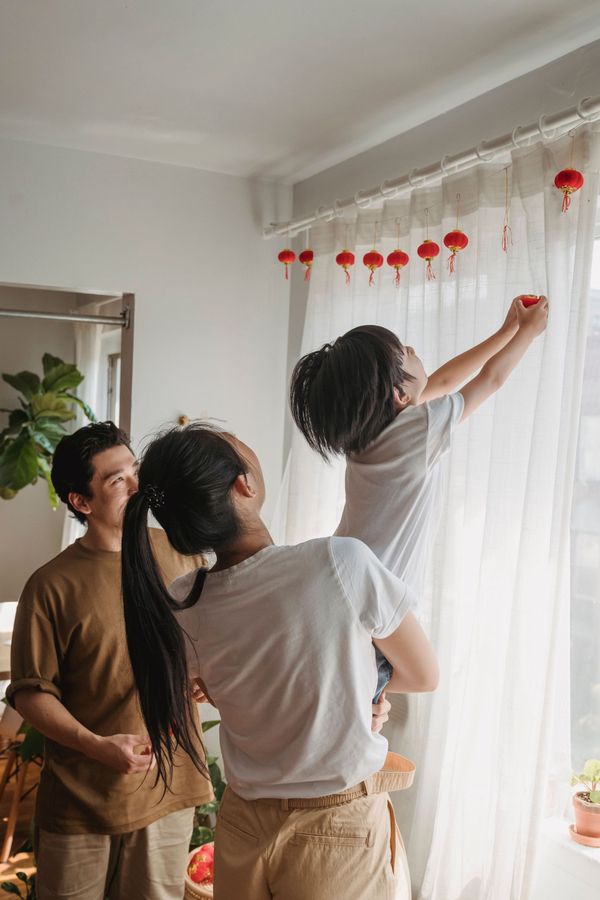 Three people hang red pom-pom garland along a sheer-curtained window.