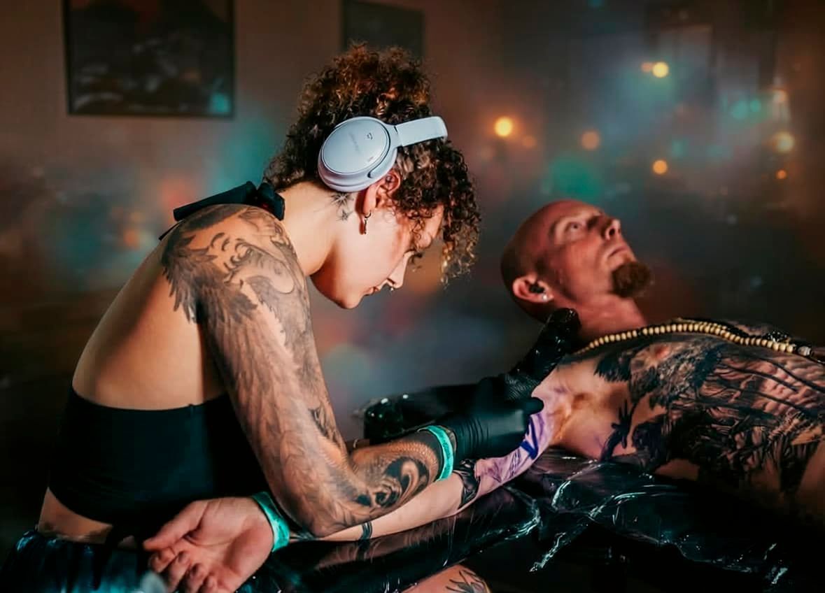 Tattoo artist working on a client in a dim, colorful studio, with headphones and ink tools visible