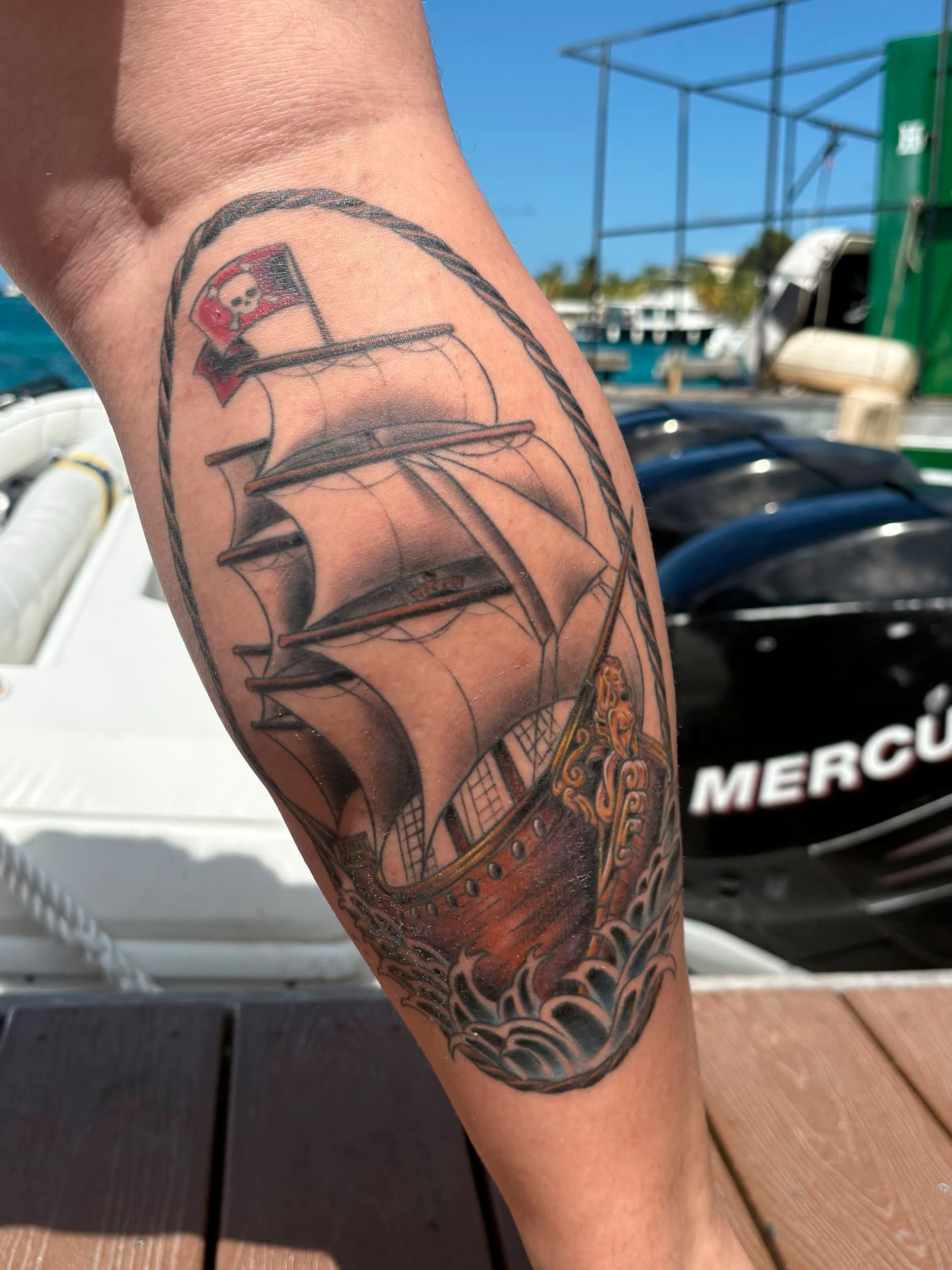 Forearm tattoo of a sailing ship with dark sails and a red-and-blue flag, outdoors by a docked boat.