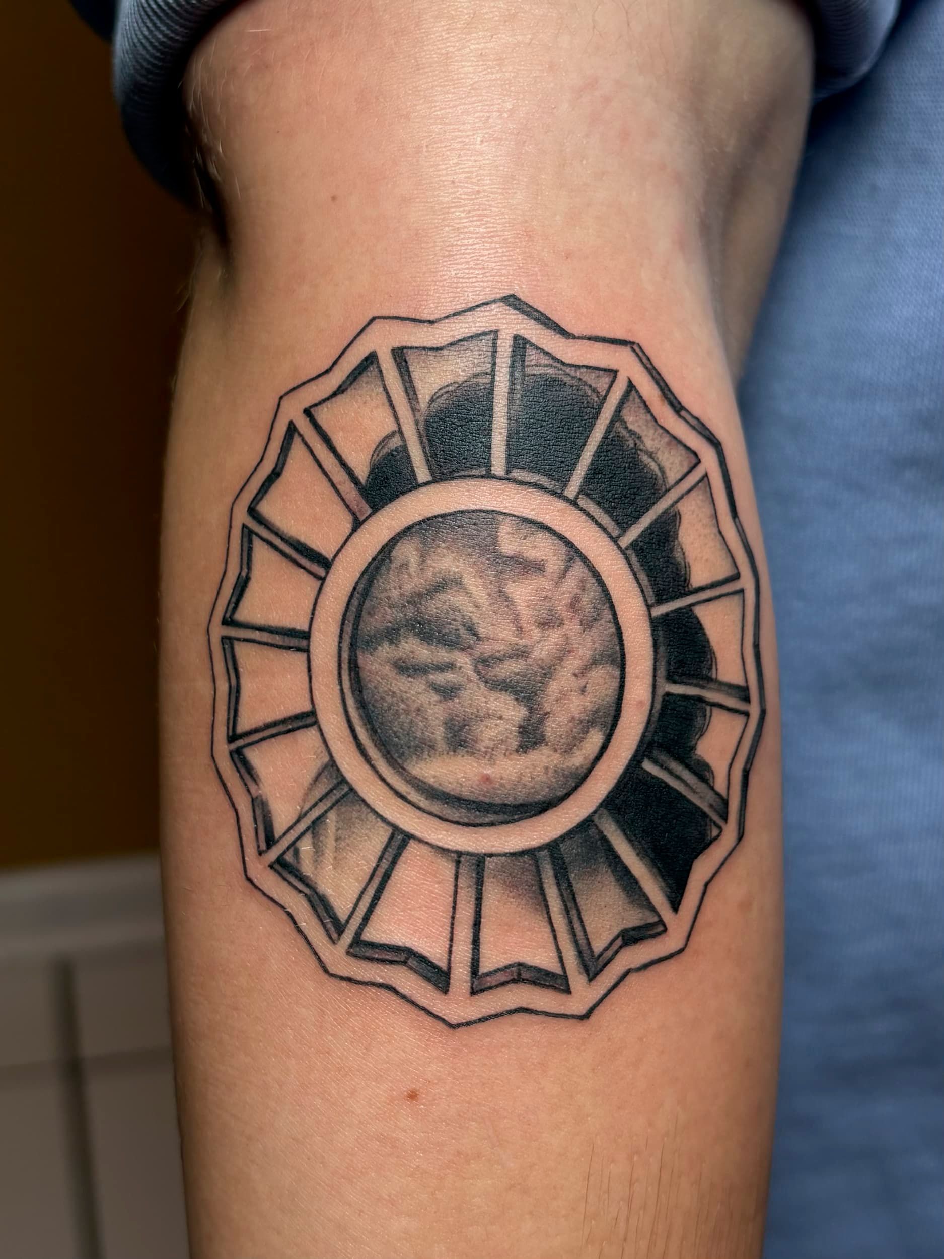 Geometric black-and-gray tattoo on a forearm, featuring a circular face at the center and radiating petal-like shapes