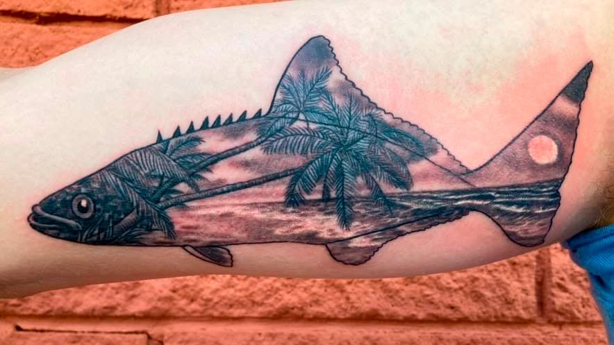 Black fish tattoo with a translucent wing-like overlay on a person’s forearm against a brick background