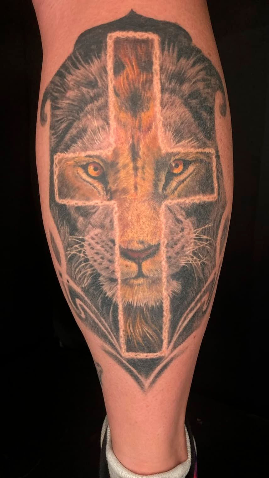 Lion face tattoo with a glowing cross on a calf, in black and orange tones.