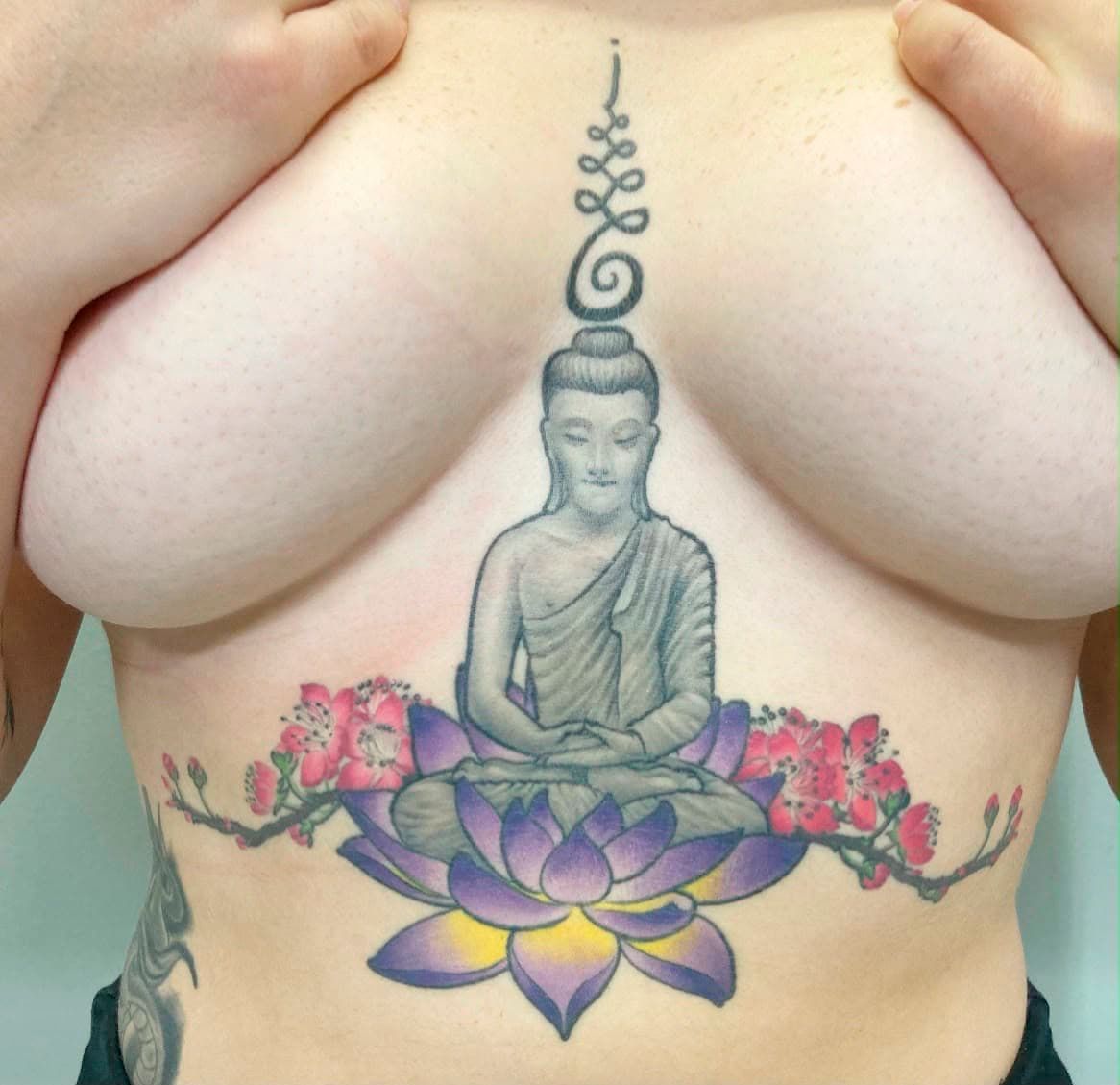 Torso tattoo of a meditating Buddha on a purple lotus with pink blossoms and decorative script above