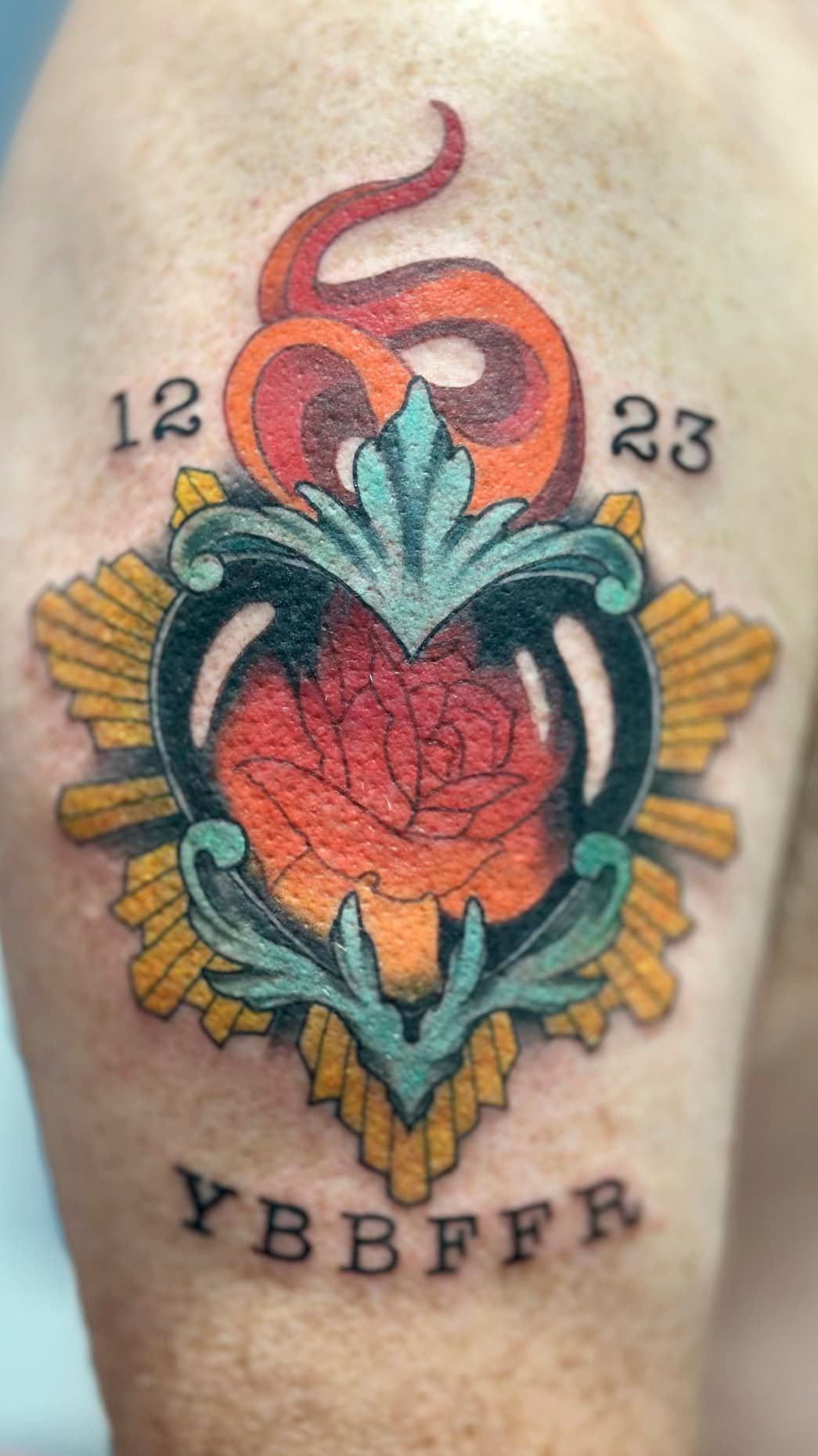 Colorful tattoo of a red heart with teal leaves, yellow rays, and dates 12, 23, plus letters YBBFRT below
