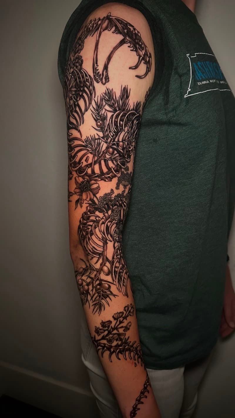 Arm in side view with a large black floral and vine tattoo on the upper arm and forearm.
