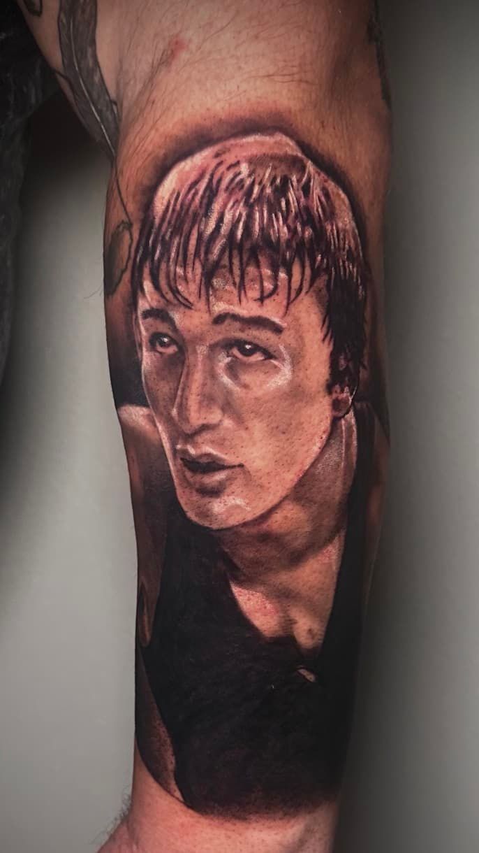 Portrait tattoo of a serious-looking face on a forearm, in black and gray ink.