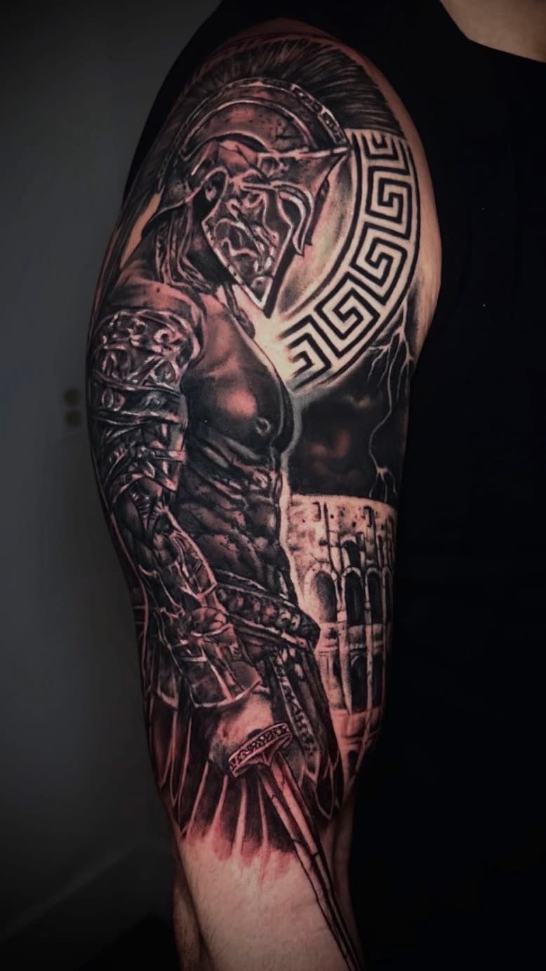 Black-and-red tattoo of a muscular warrior’s arm with a helmet and geometric patterns on dark skin