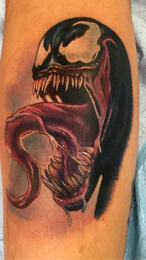 Black and red Venom-style tattoo on skin, with open mouth, long tongue, and sharp teeth.