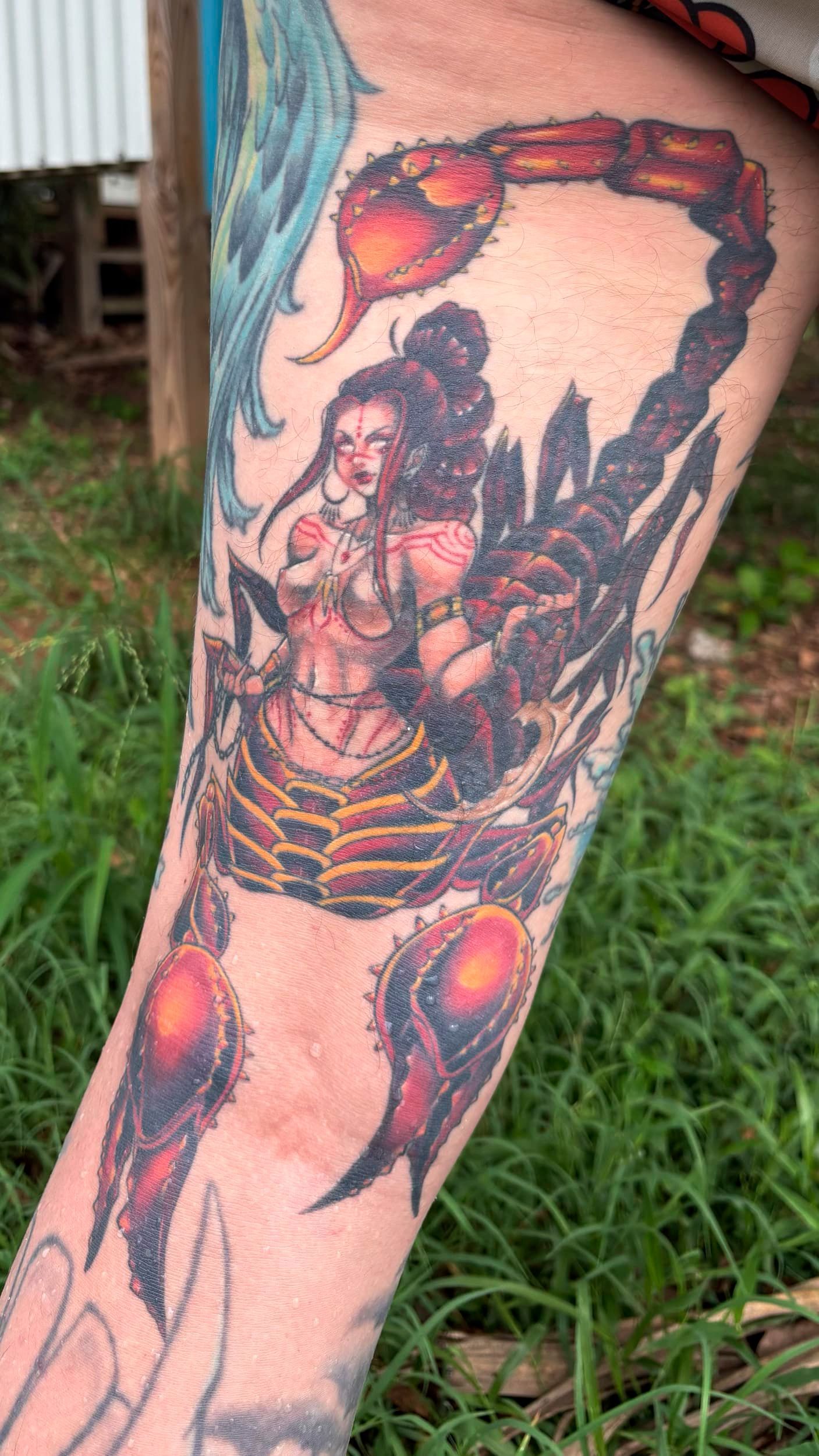 Colorful tattoo on a forearm showing a seated figure with a large scorpion-like tail and glowing red orbs
