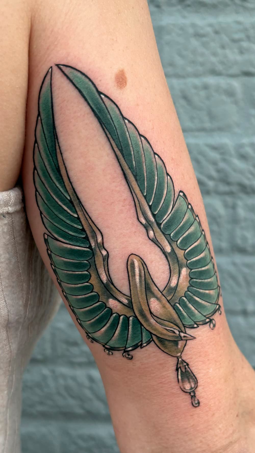 Green and pink fantasy creature tattoo on a forearm, with wing-like fins and dangling details