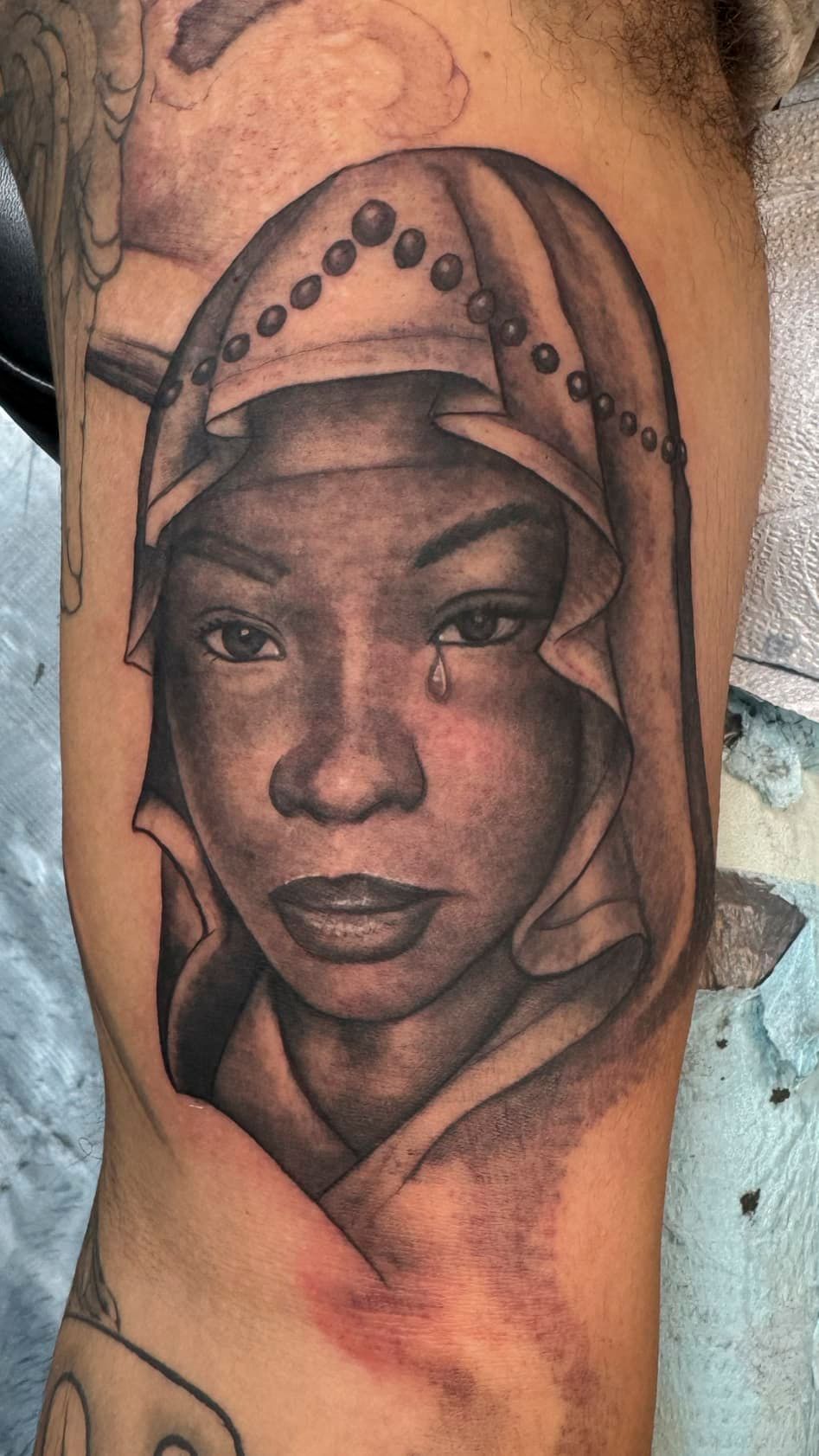 Black-and-gray tattoo portrait of a hooded face with detailed shading on an arm.