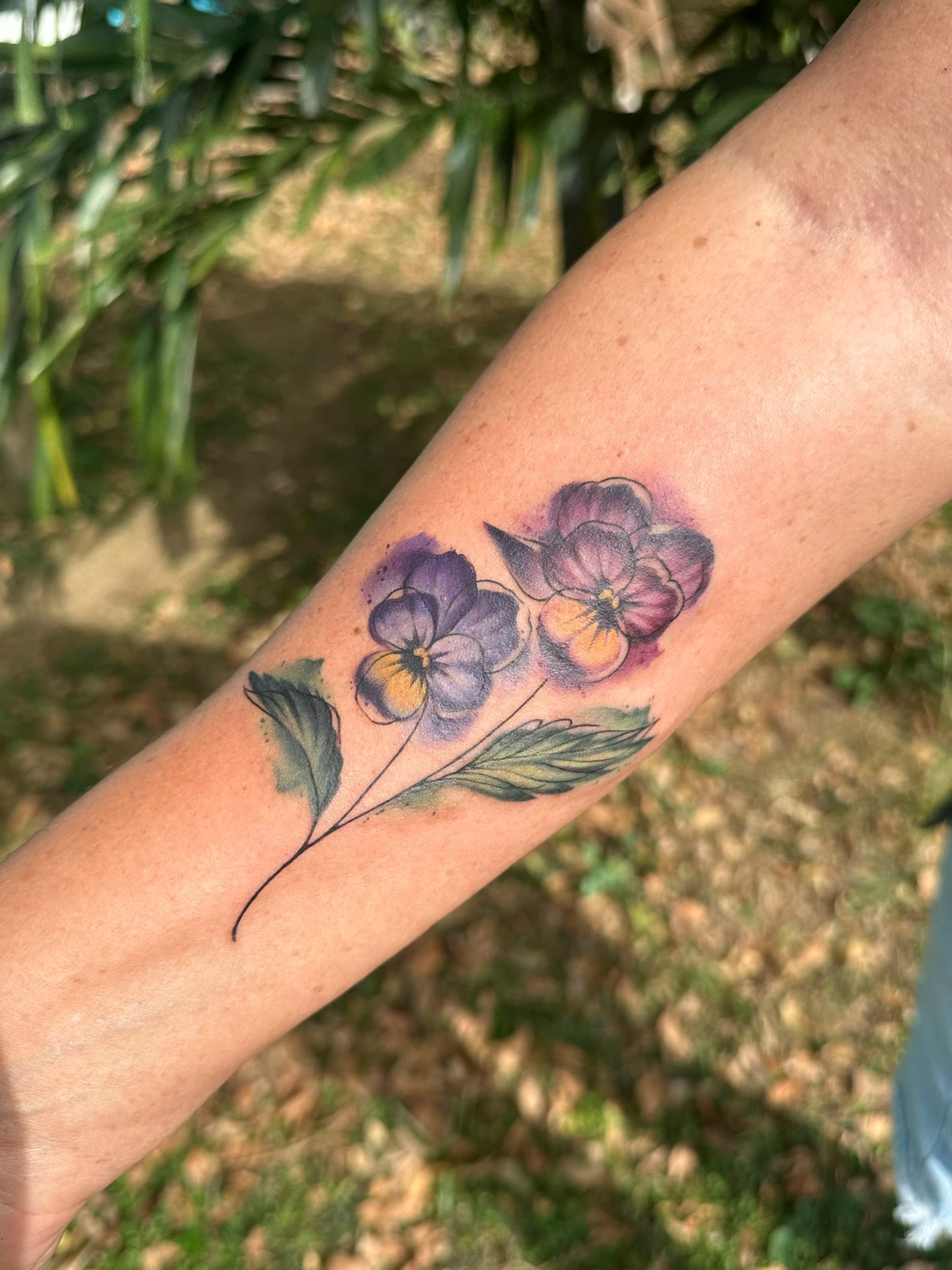 Forearm tattoo of purple flowers with green leaves outdoors in sunlight