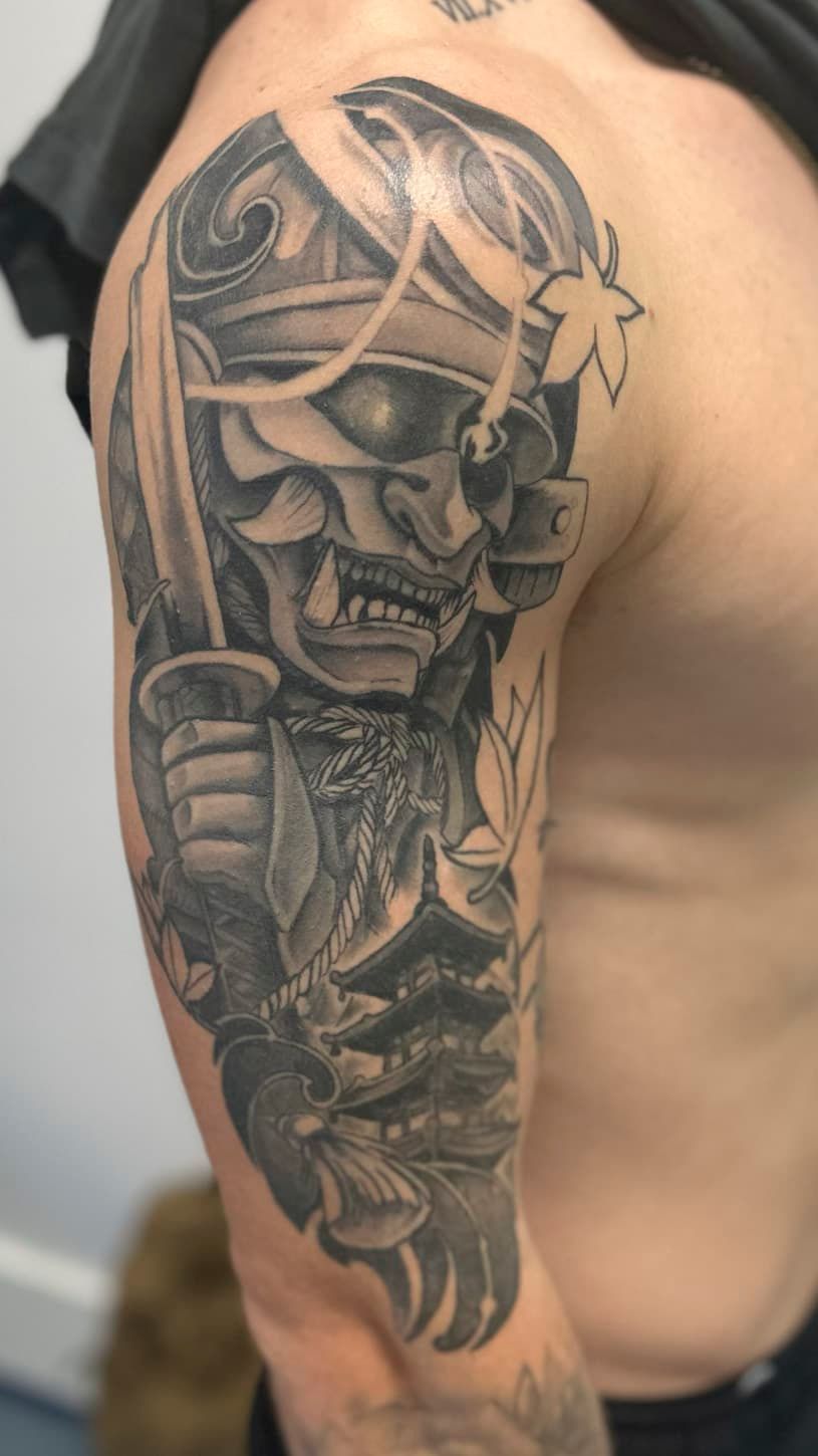Black-and-gray tattoo of a skull-like warrior face with floral details on an upper arm