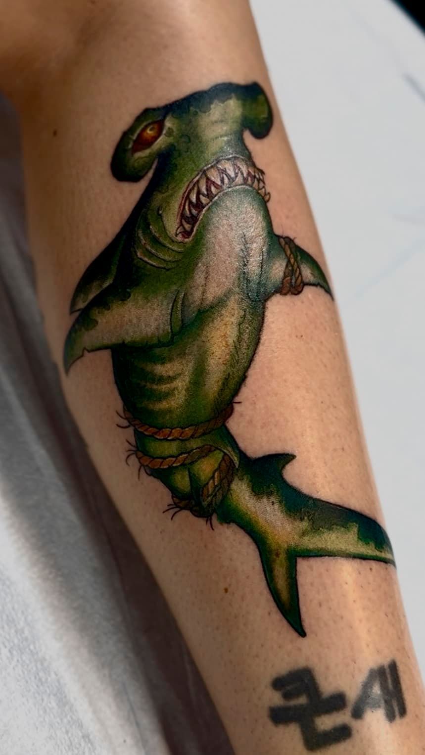 Tattoo of a green hammerhead shark on a forearm, with a small black symbol near the wrist