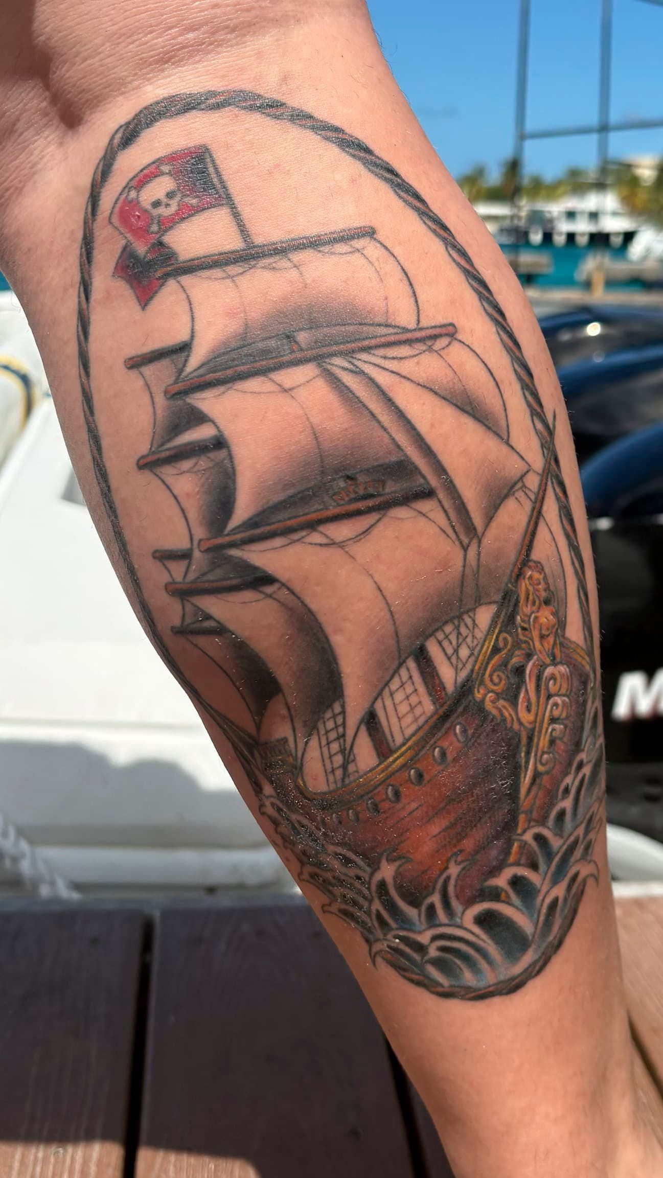 Black-and-gray tattoo of a sailing ship on a forearm, with a skull and rope details near the wrist.