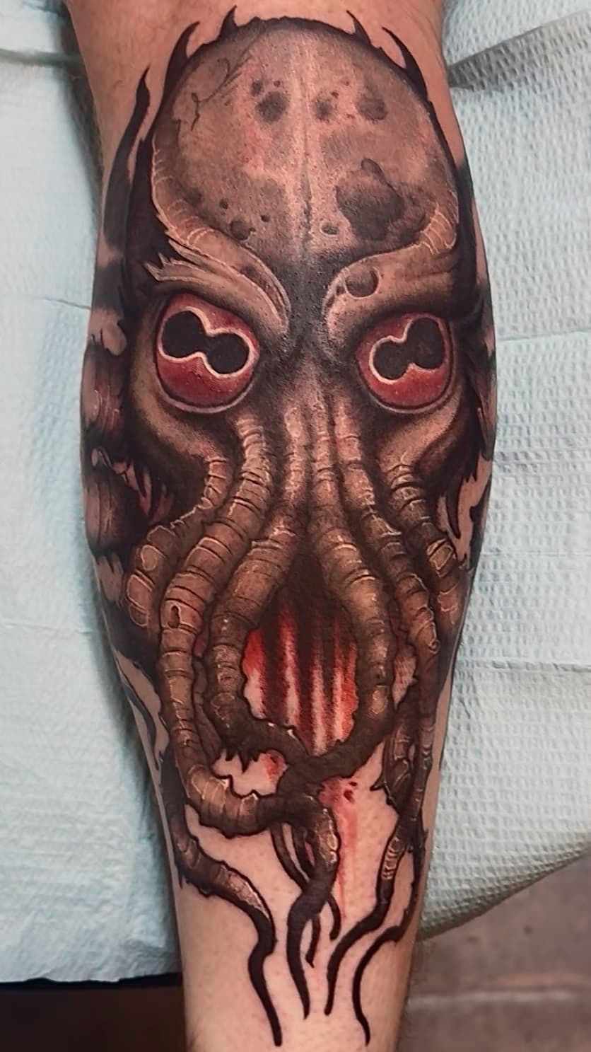 Dark tattoo of a tentacled Cthulhu-like face with glowing red eyes on a forearm