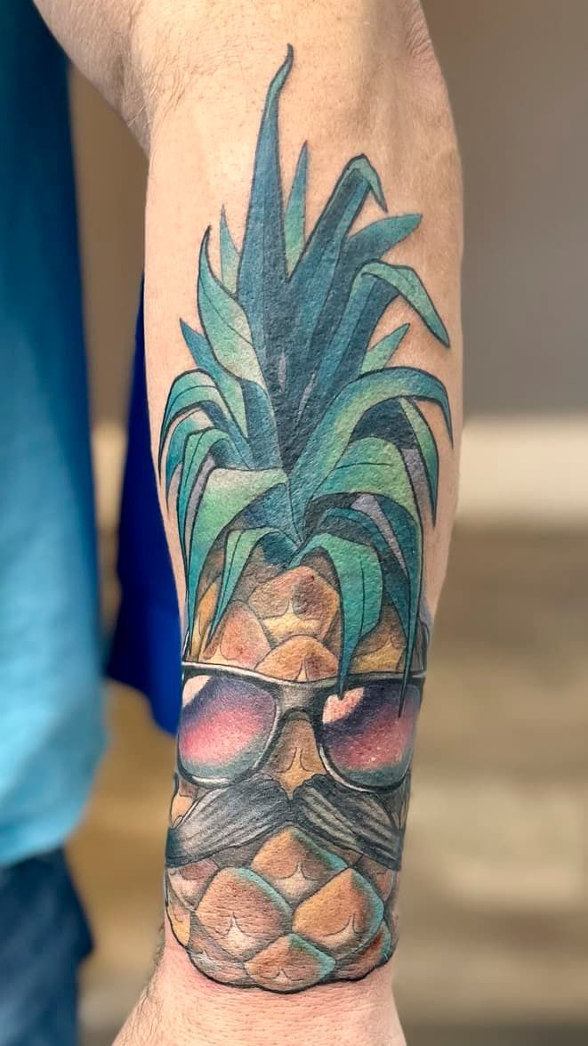 Colorful pineapple tattoo on a person’s forearm, with teal leaves and warm brown fruit tones.