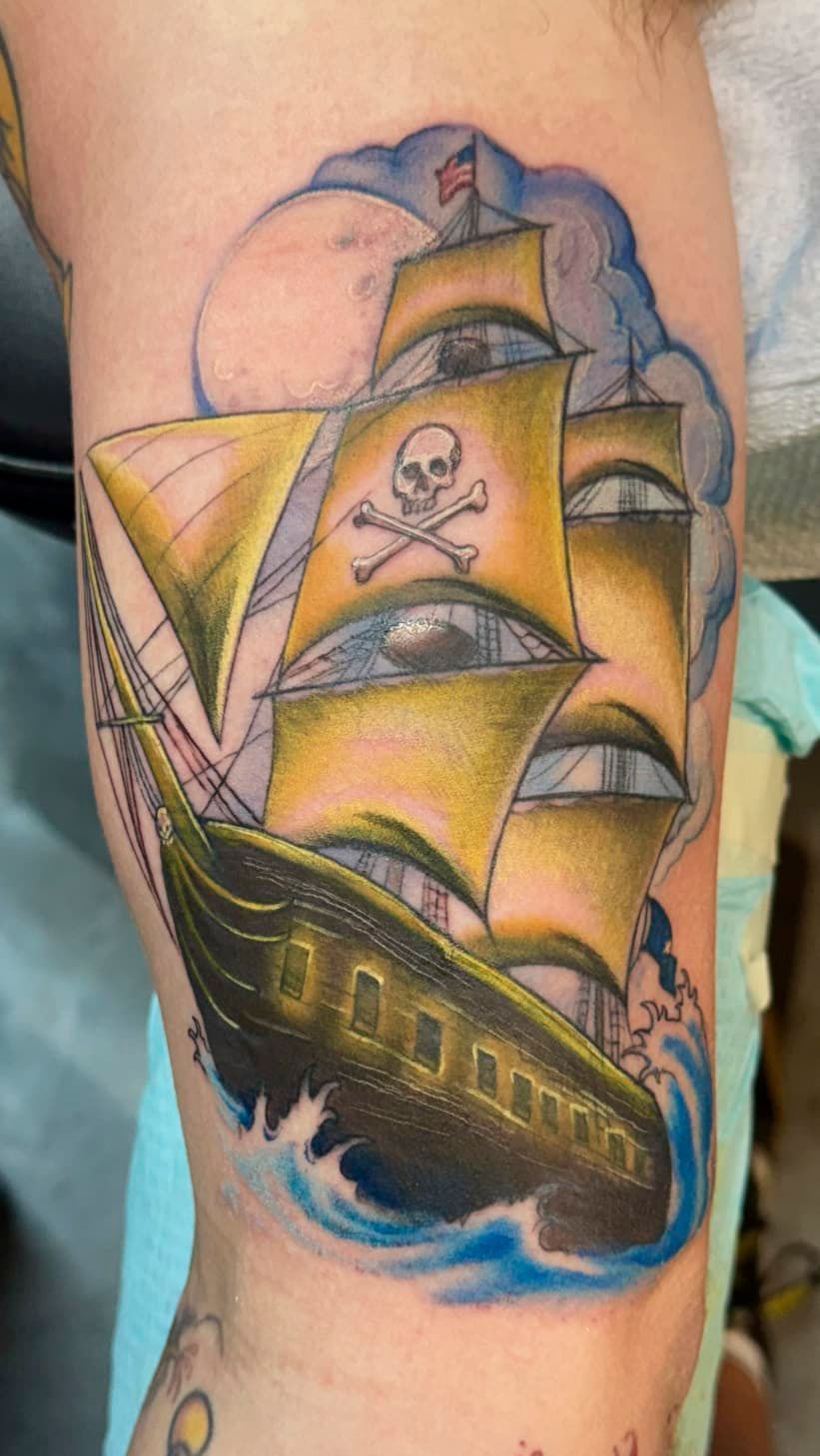 Tattoo of a sailing ship with skull-and-crossbones sails in blue, yellow, and black on skin