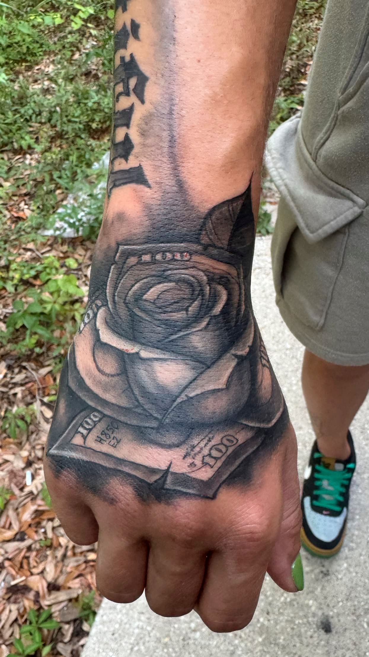 Tattooed hand and forearm with a black rose design, shown outdoors near a sidewalk and grass.