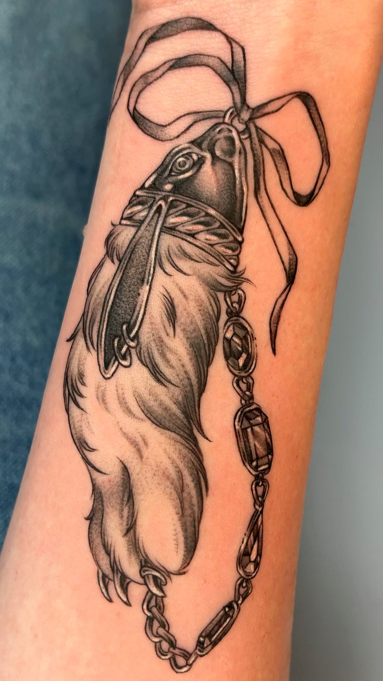 Black-and-gray tattoo of a feather tied with a ribbon and beaded chain on skin