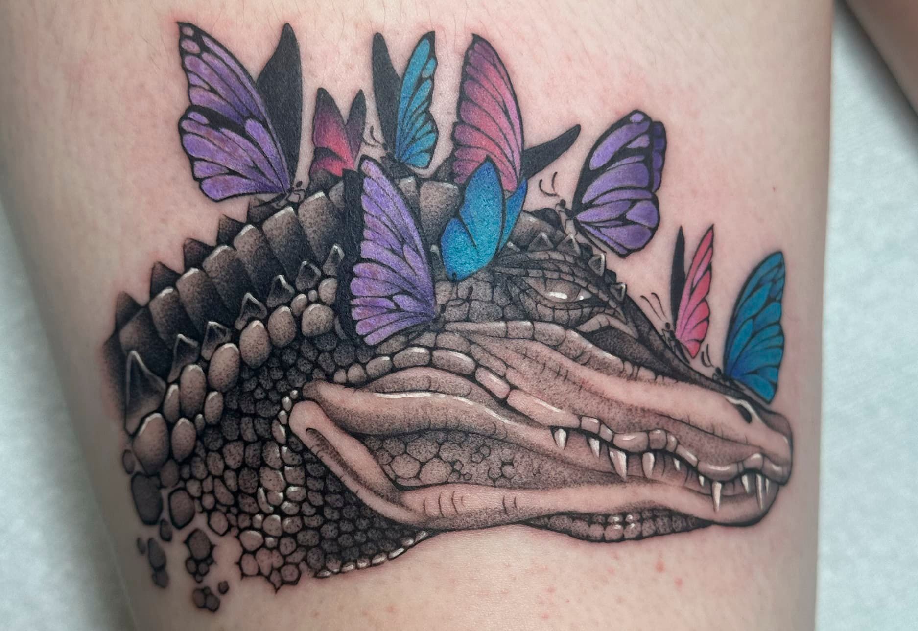 Close-up tattoo of a reptile head with purple and blue butterflies on top, in black and gray shading