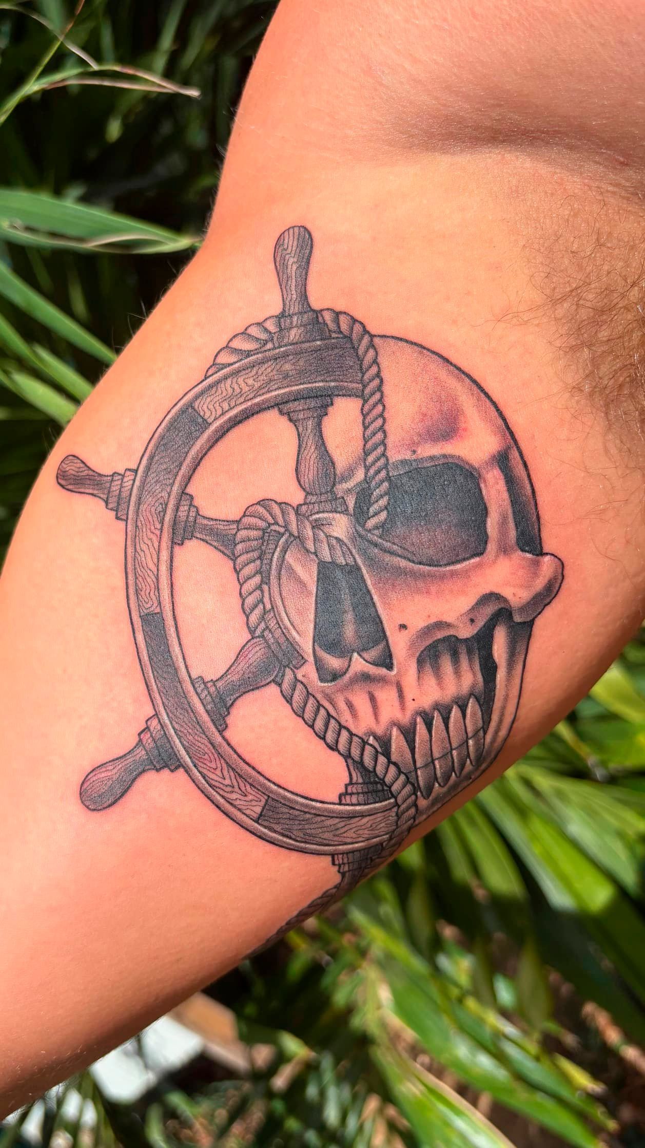 Black compass-style tattoo on a person's upper arm outdoors, with green foliage in the background