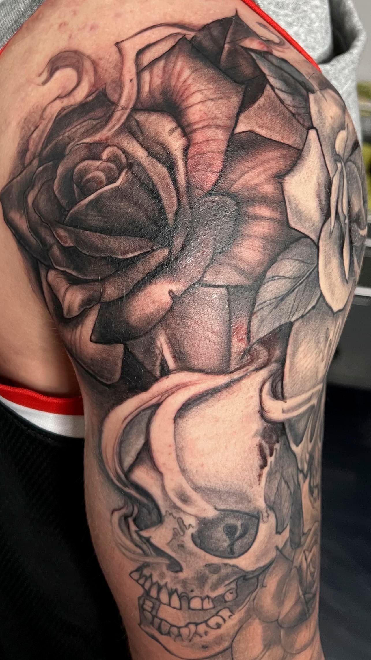 Black-and-gray rose and skull tattoo on an upper arm/shoulder with red fabric strap nearby