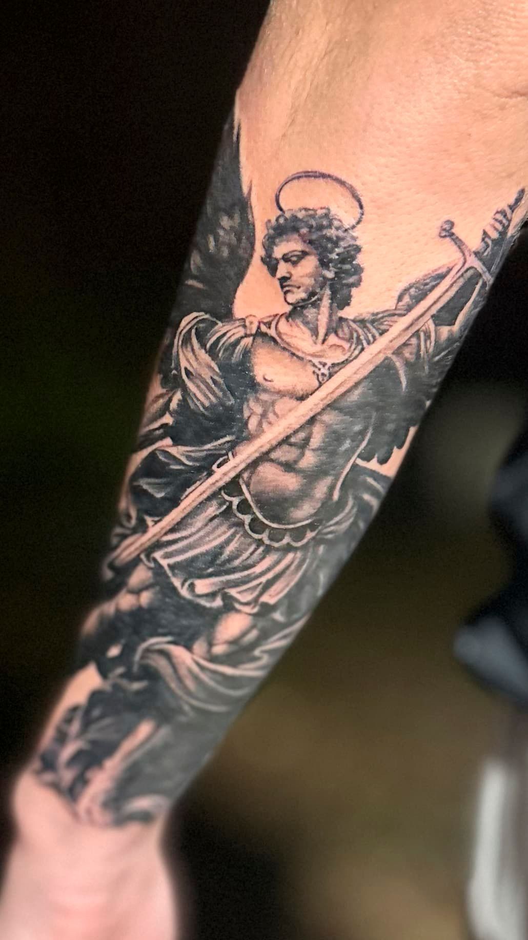 Black and gray tattoo of a haloed robed figure on a forearm